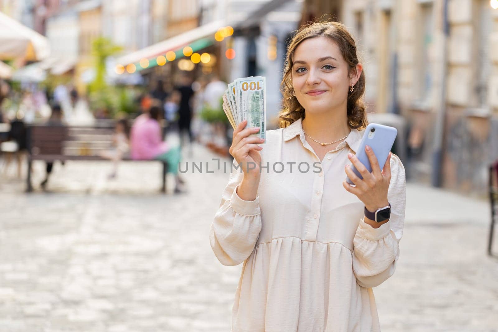 Happy young woman counting money dollar cash, use smartphone calculator app, satisfied of income for planned vacation gifts. Lovely girl walking in urban city sunny street. Town lifestyles outdoors