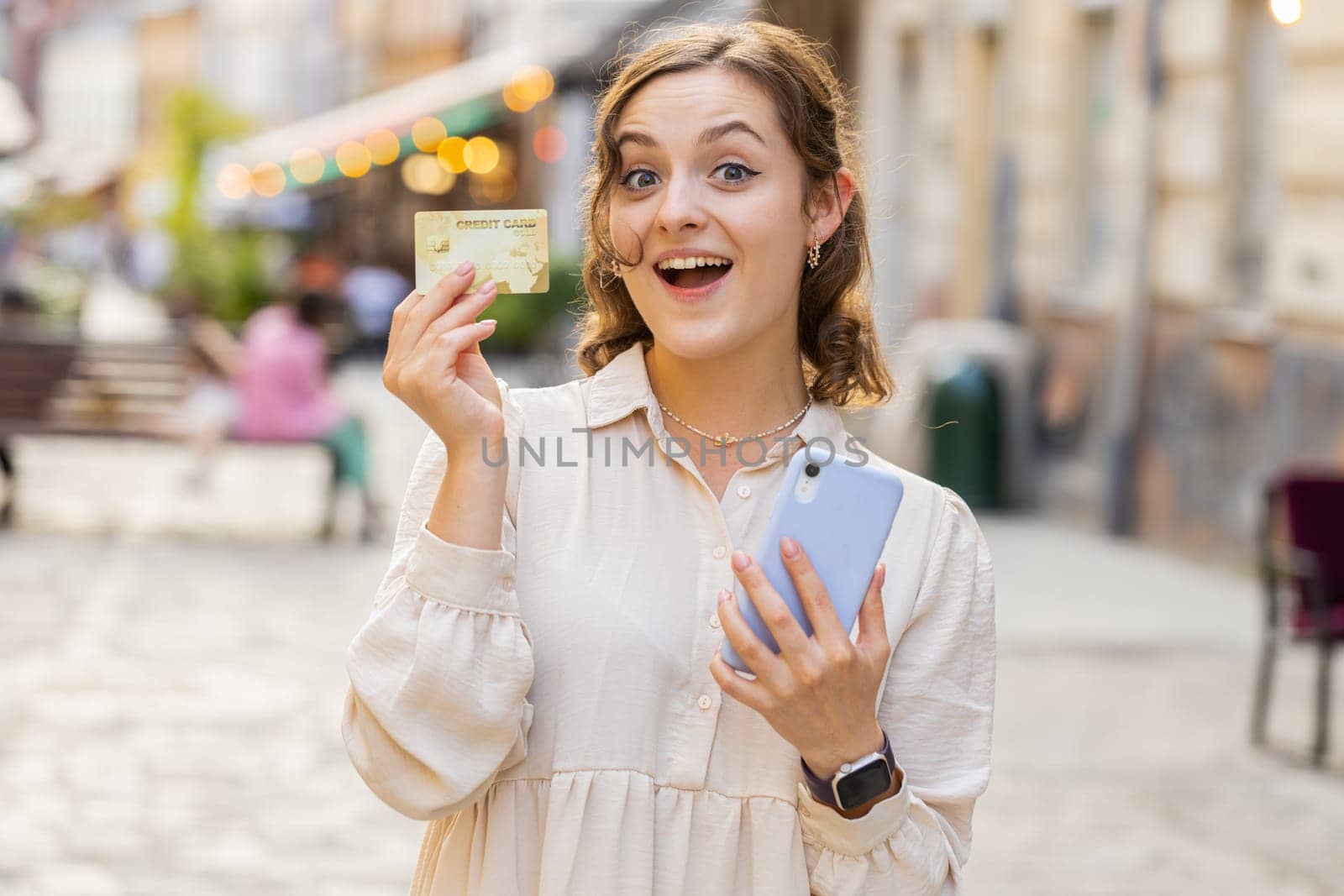 Young woman using credit bank card smartphone while transferring money, purchases online shopping, order food delivery, booking hotel room. Lovely girl walking in urban city sunny street outdoors