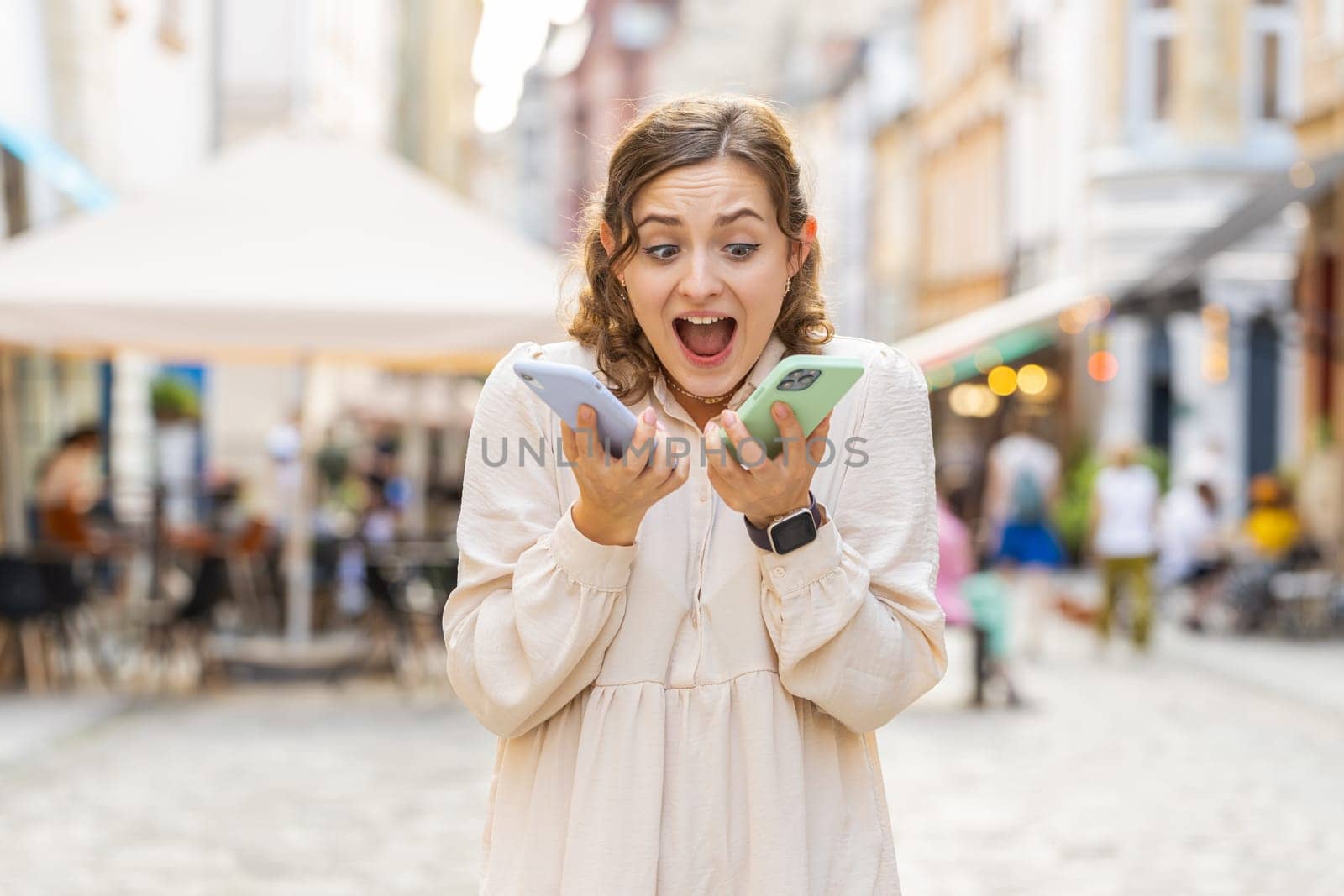 Irritated nervous stressed young woman talking screaming on two mobile phones having conversation conflict quarrel complaint dispute discuss solve problem outdoors. Girl walking in urban city street