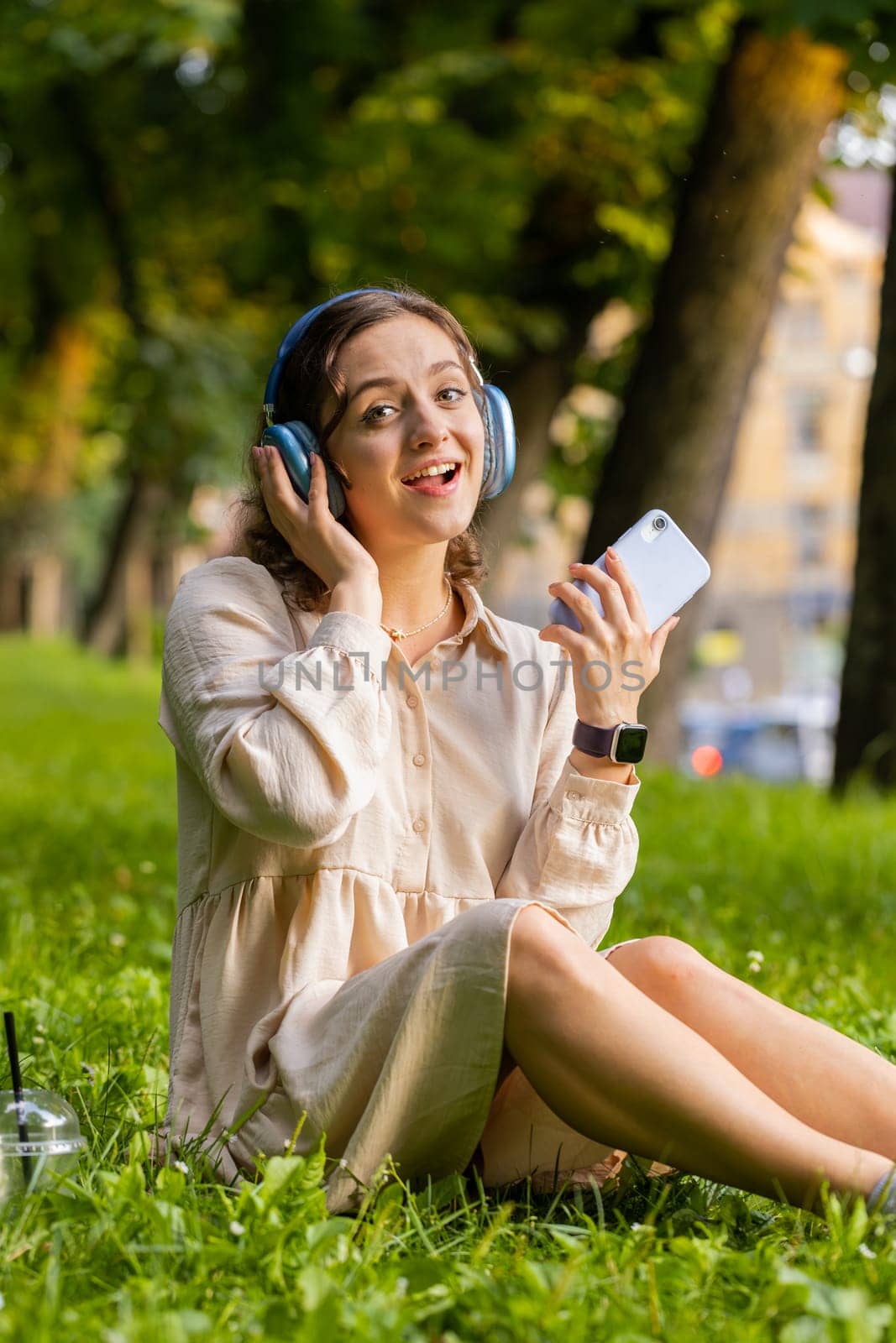 Happy relaxed young woman in wireless headphones choosing, listening favorite energetic disco rock n roll music in smartphone dancing outdoor. Girl sitting on grass in urban sunset city park. Vertical