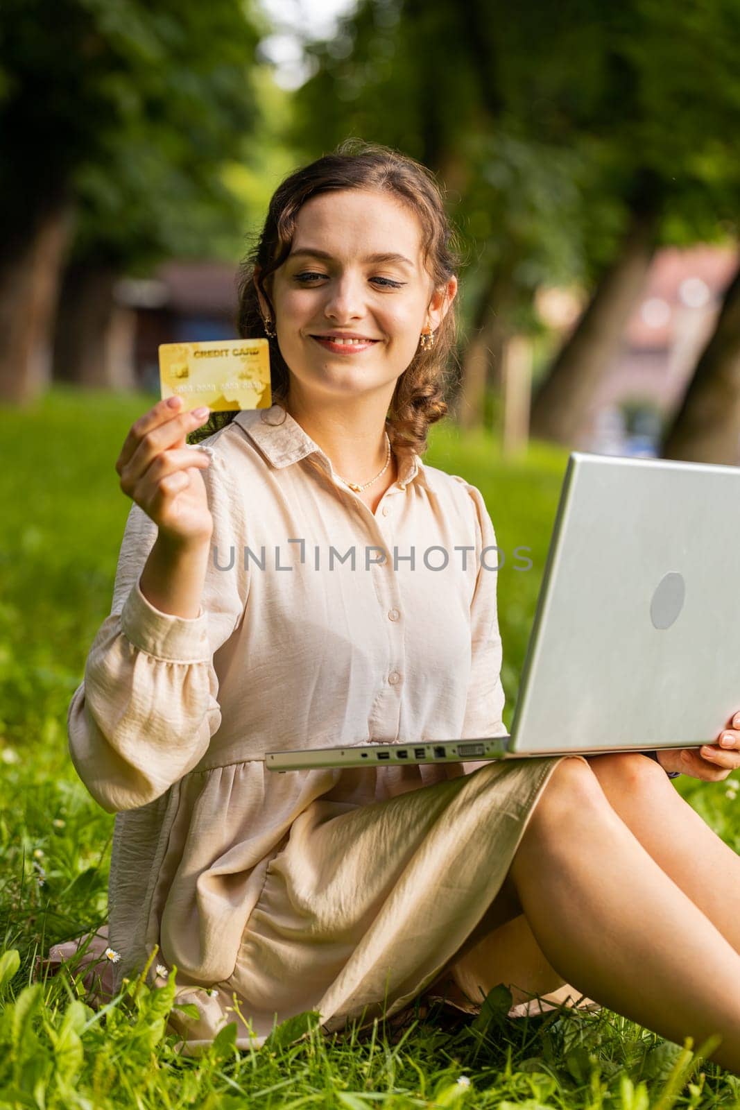 Young woman using credit bank card and laptop computer while transferring money, purchases online shopping, order food delivery. Girl sitting on grass in urban sunset city park background. Vertical