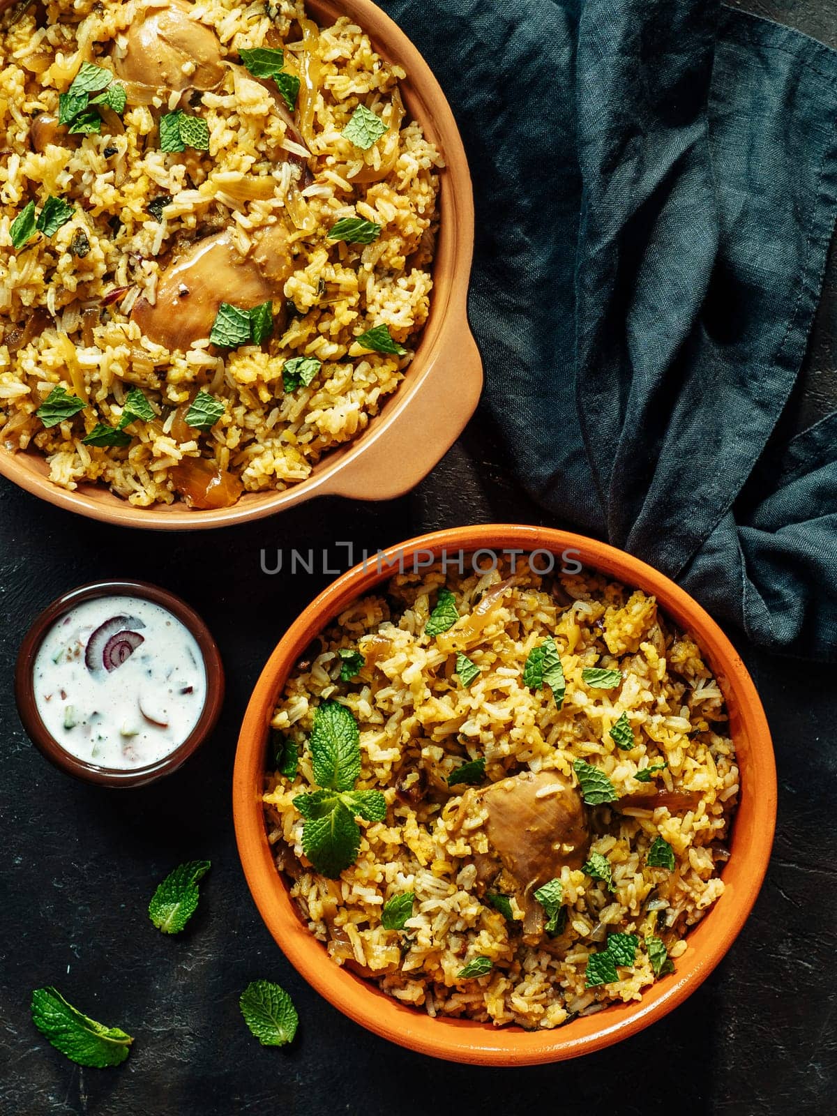 Pakistani food - biryani rice with chicken and raita yoghurt dip. Delicious hyberabadi chicken biryani in two bowl over black background. Top view or flat lay. Vertical.