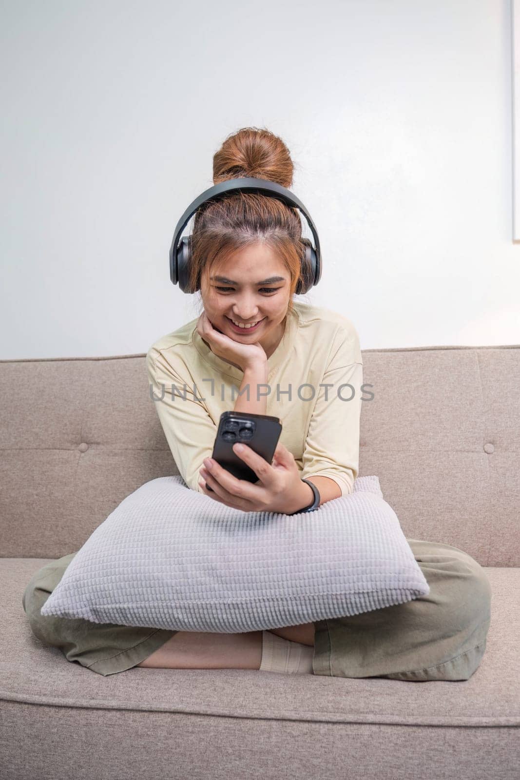 A charming young Asian woman in casual clothes enjoys the music on her headphones while sitting on a sofa in her living room. by wichayada