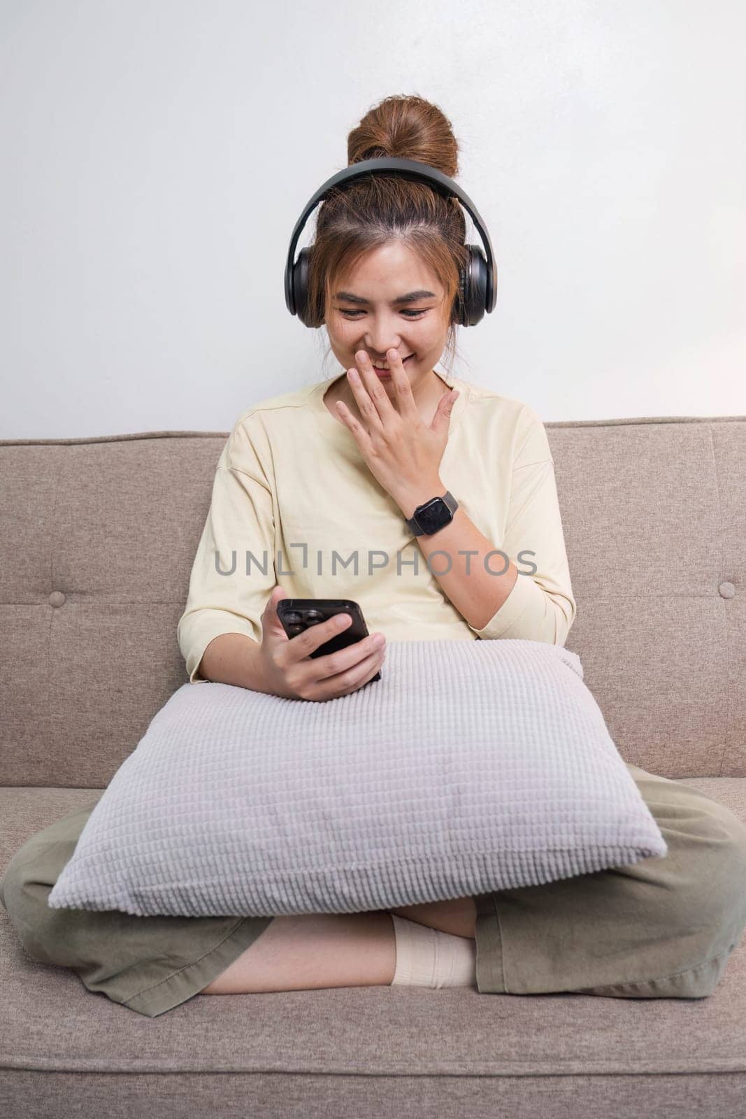 A charming young Asian woman in casual clothes enjoys the music on her headphones while sitting on a sofa in her living room...