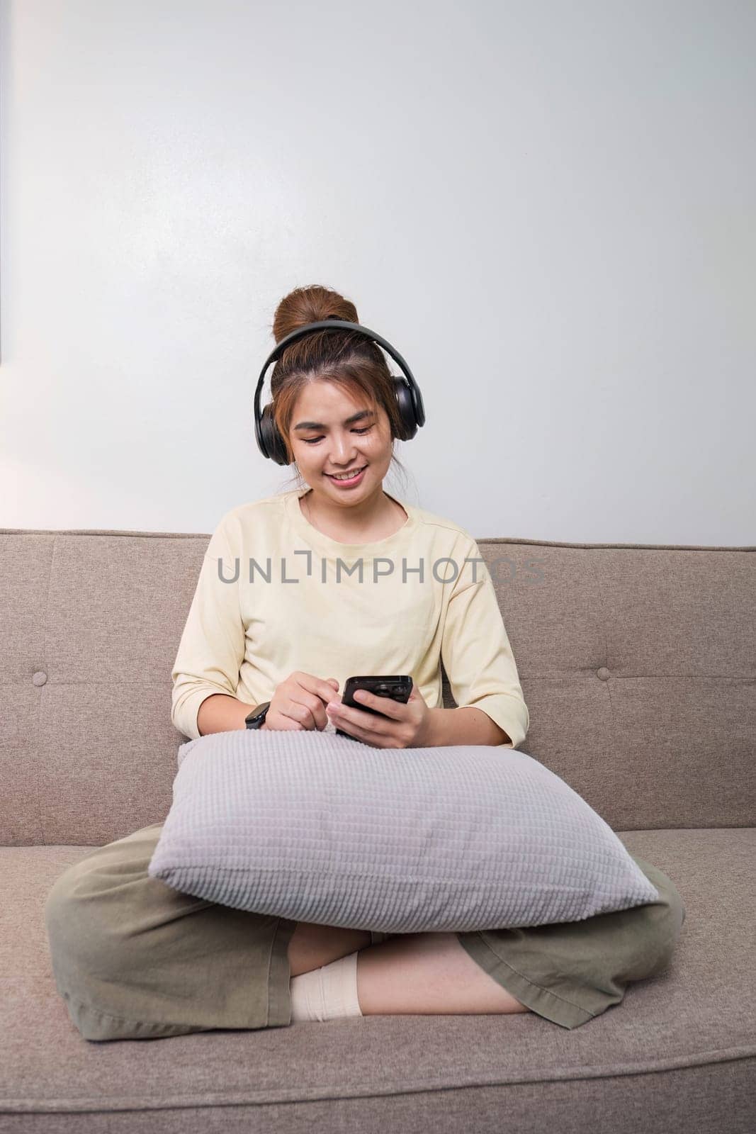 A charming young Asian woman in casual clothes enjoys the music on her headphones while sitting on a sofa in her living room...