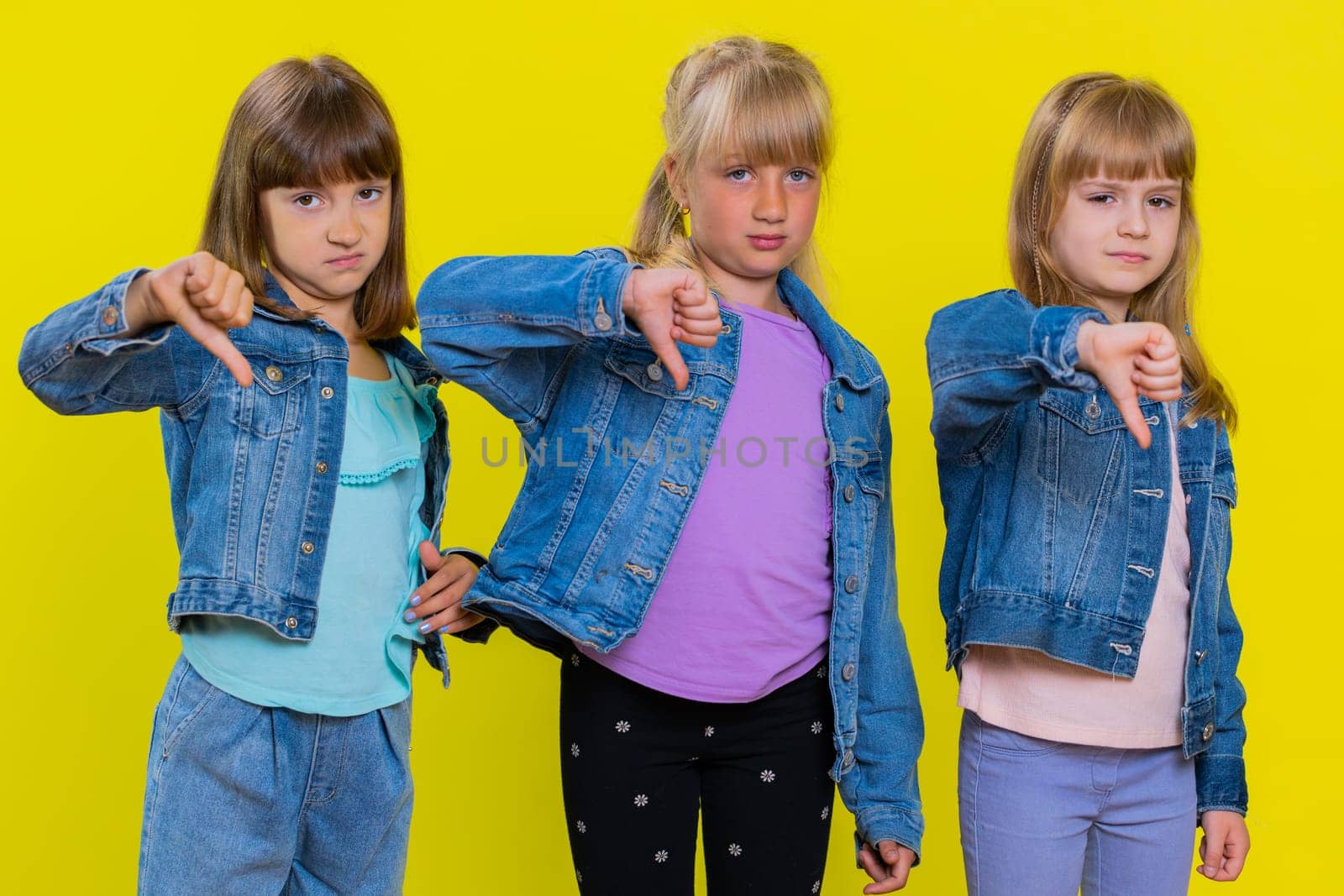 Dislike. Upset teenage girls showing thumbs down sign gesture, expressing discontent, disapproval, dissatisfied. Little children sisters. Three cute siblings kids isolated on studio yellow background