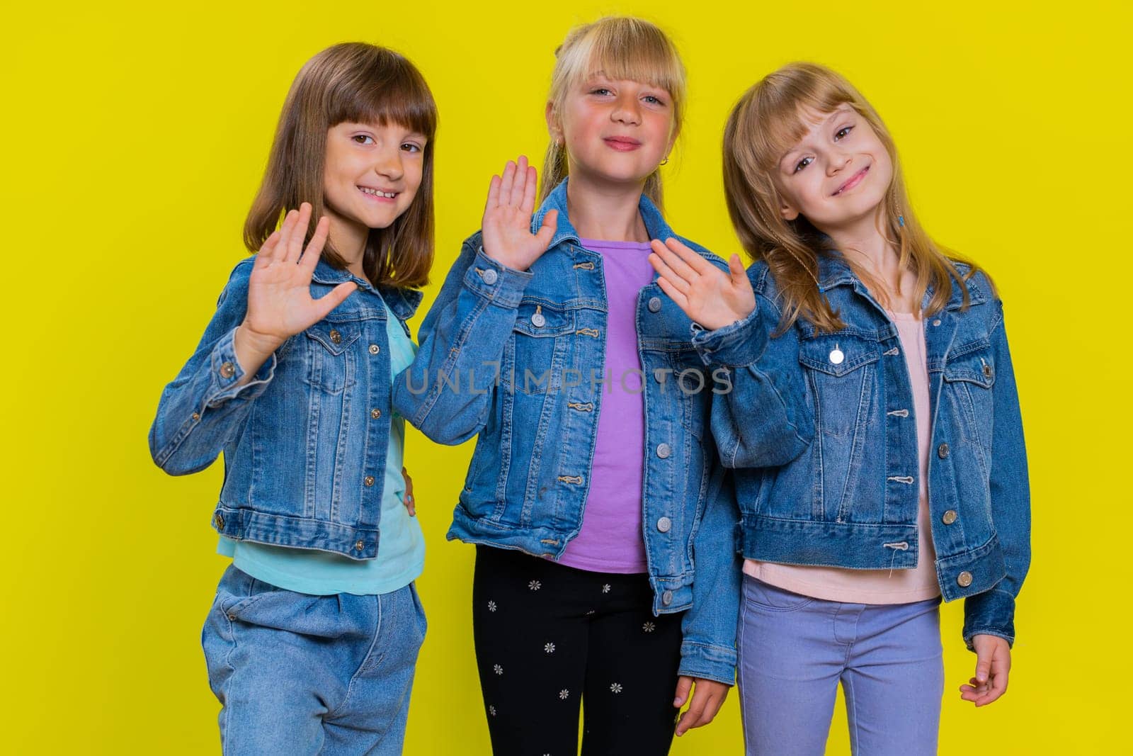 Teenage girls smiling friendly at camera and waving hands gesturing hello or goodbye, welcoming with hospitable expression. Little children sisters. Three siblings kids on studio yellow background