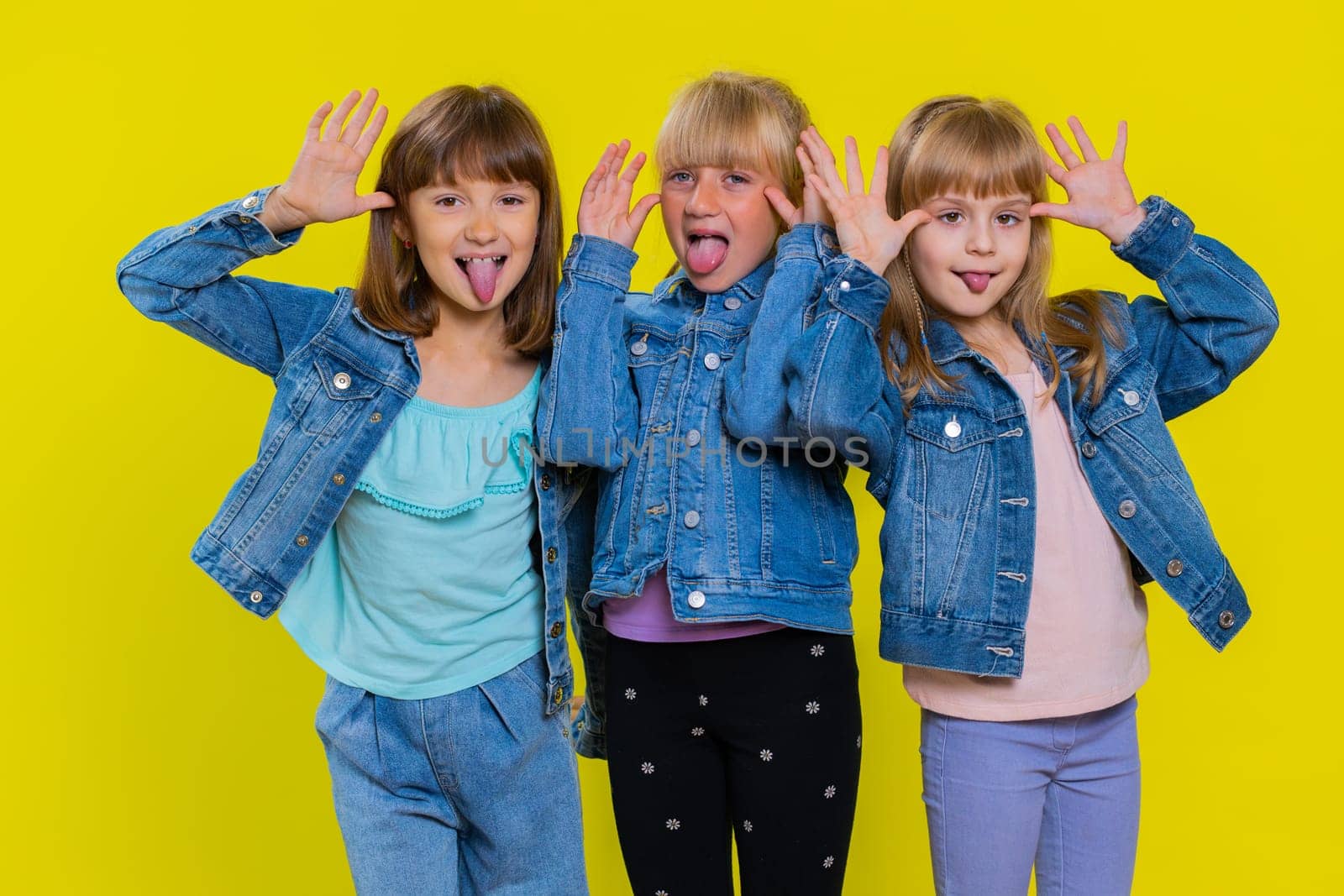 Funny happy teenage girls making playful silly facial expressions and grimacing, fooling around, showing tongue. Little children sisters. Three cute siblings kids isolated on studio yellow background