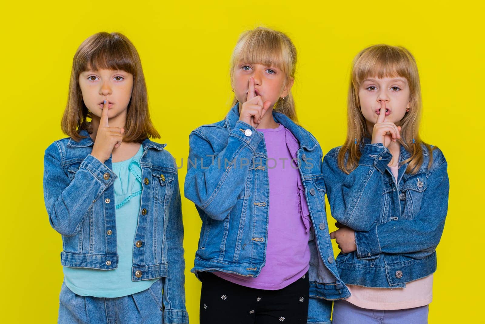 Shh be quiet please. Teenage cute girls presses index finger to lips makes silence gesture sign do not tells secret. Little children sisters. Three siblings kids isolated on studio yellow background