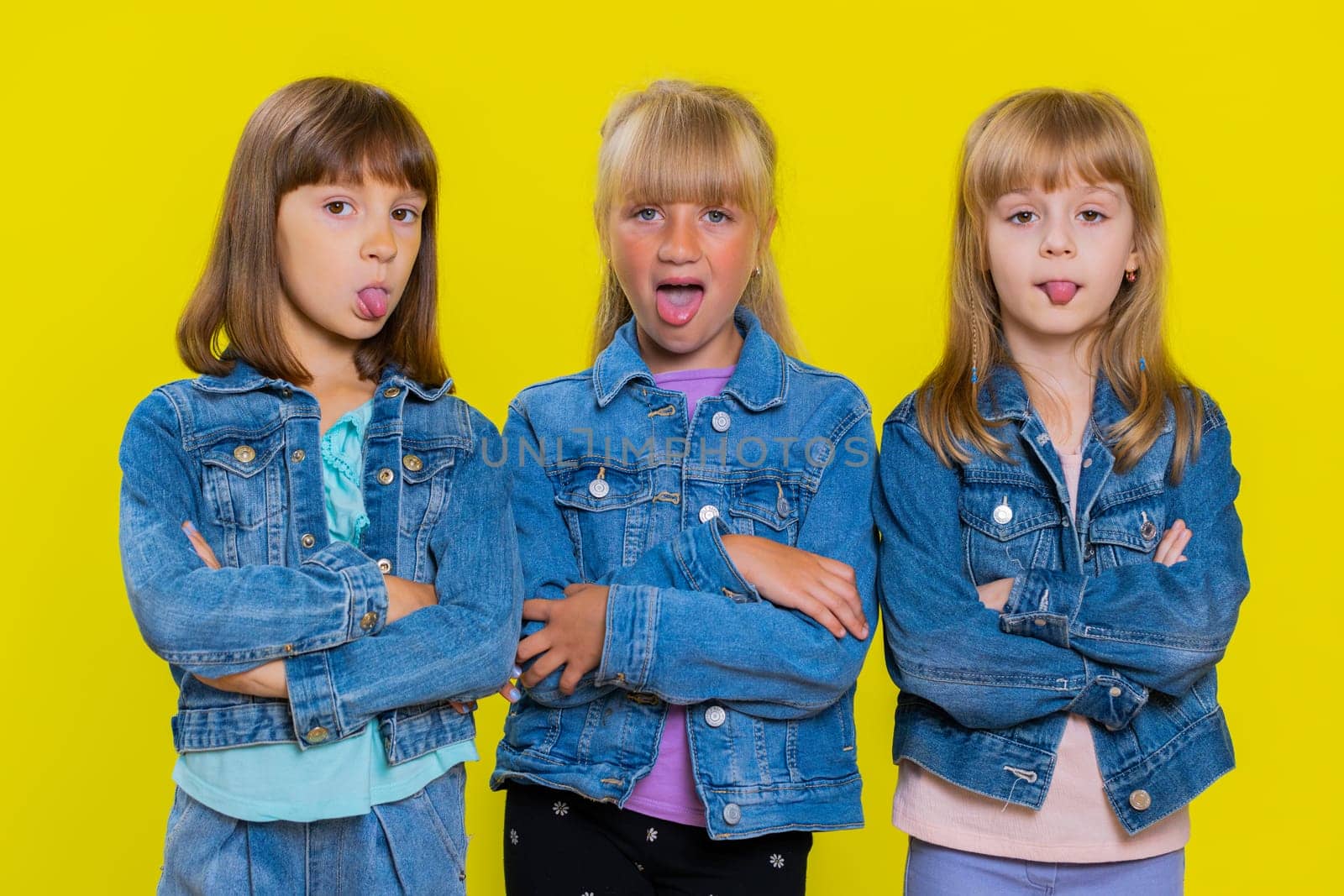 Teenage girls showing tongue making faces at camera, fooling around, joking, aping with silly face, teasing, bullying, abuse. Little children sisters. Three siblings kids isolated on yellow background
