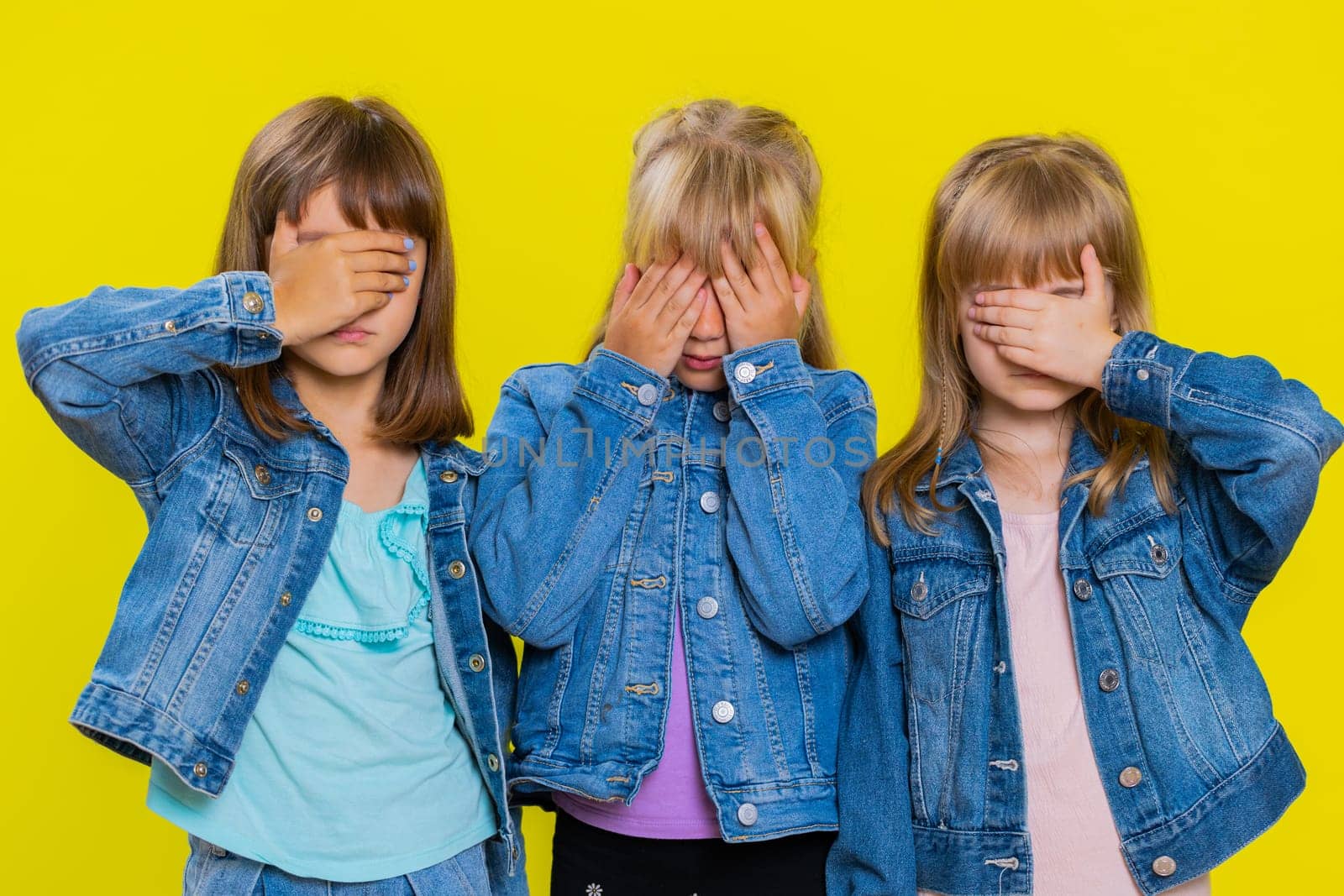Dont want to look at this, awful. Teenage girls closing eyes with hand showing stop gesture, confused shy scared to watch. Little children sisters. Three siblings kids isolated on yellow background