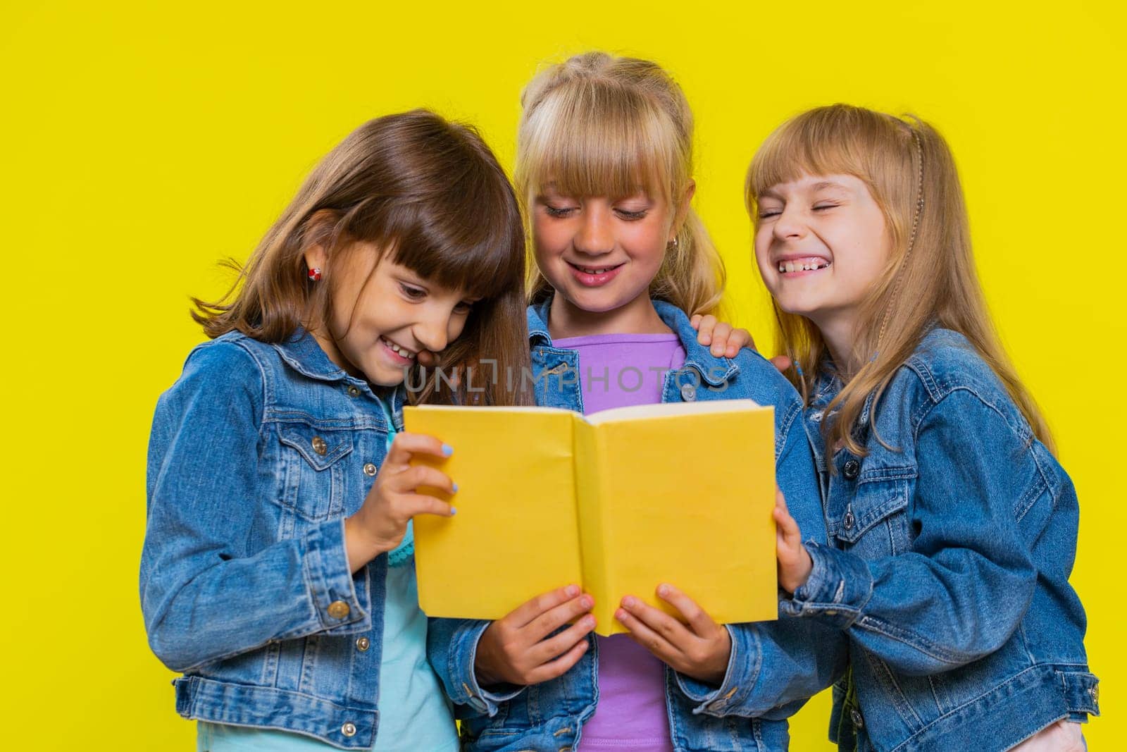 Teenage girls reading funny interesting fairytale story book, leisure hobby, knowledge wisdom, education, learning, study, wow. Little children sisters. Three cute siblings kids on yellow background