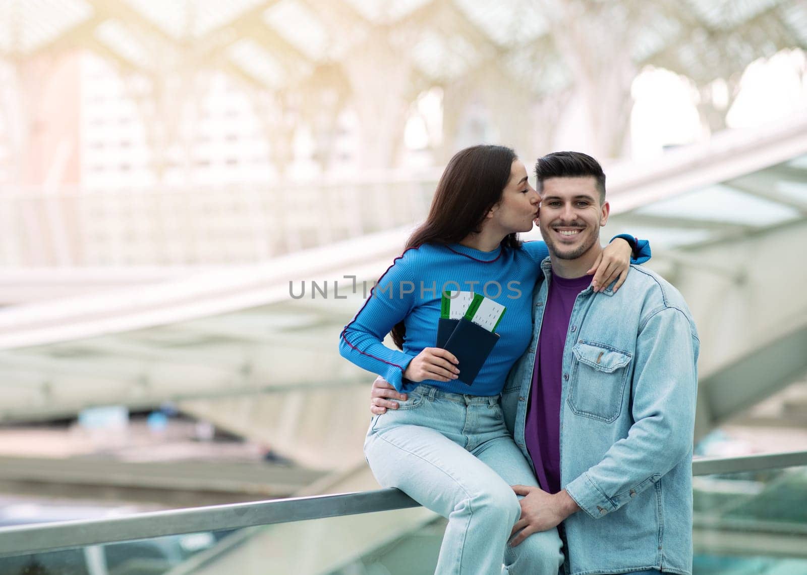 Great Travel Offer. Joyful Woman Kissing Her Husband Holding Passports With Boarding Pass Tickets Posing In Modern Airport Indoors. Couple Celebrating Vacation. Empty Space For Text