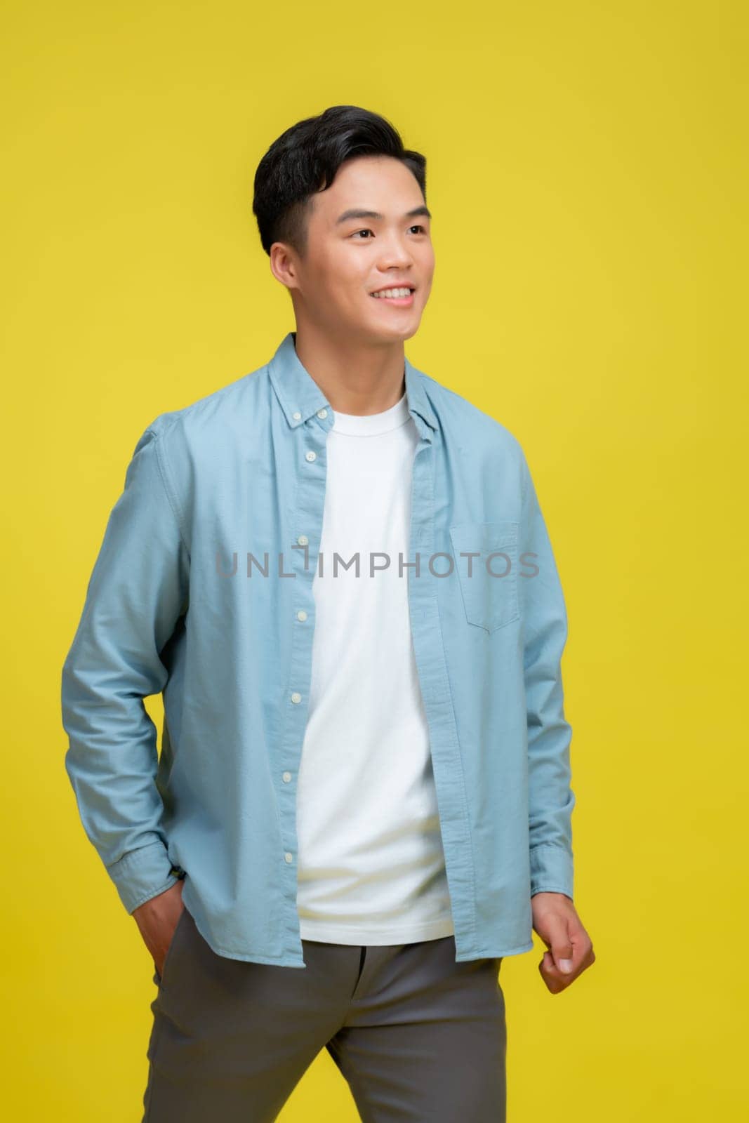  smiling young handsome Asian man standing with one hand in pocket isolated on yellow studio background