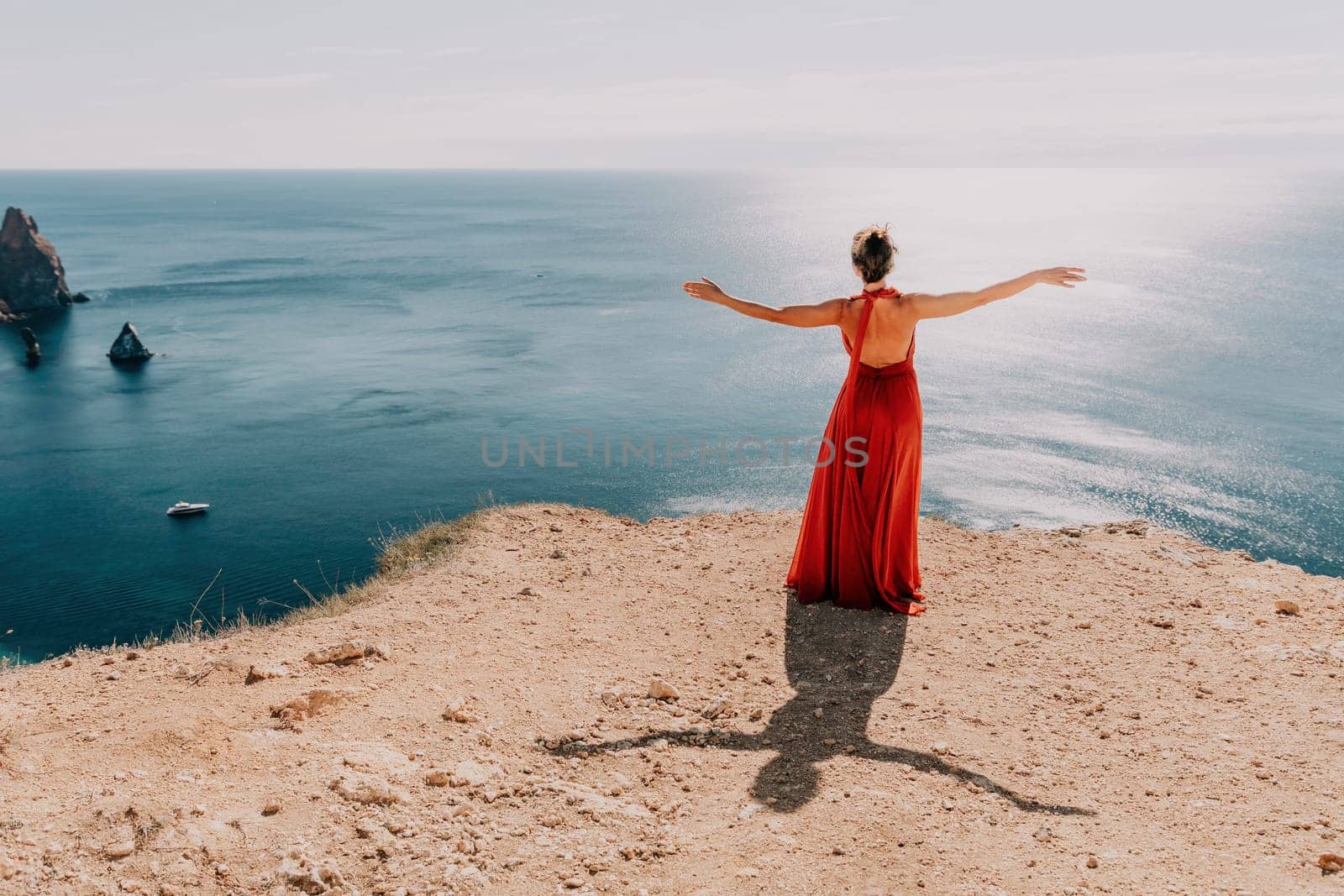 Side view a Young beautiful sensual woman in a red long dress posing on a rock high above the sea during sunrise. Girl on the nature on blue sky background. Fashion photo.