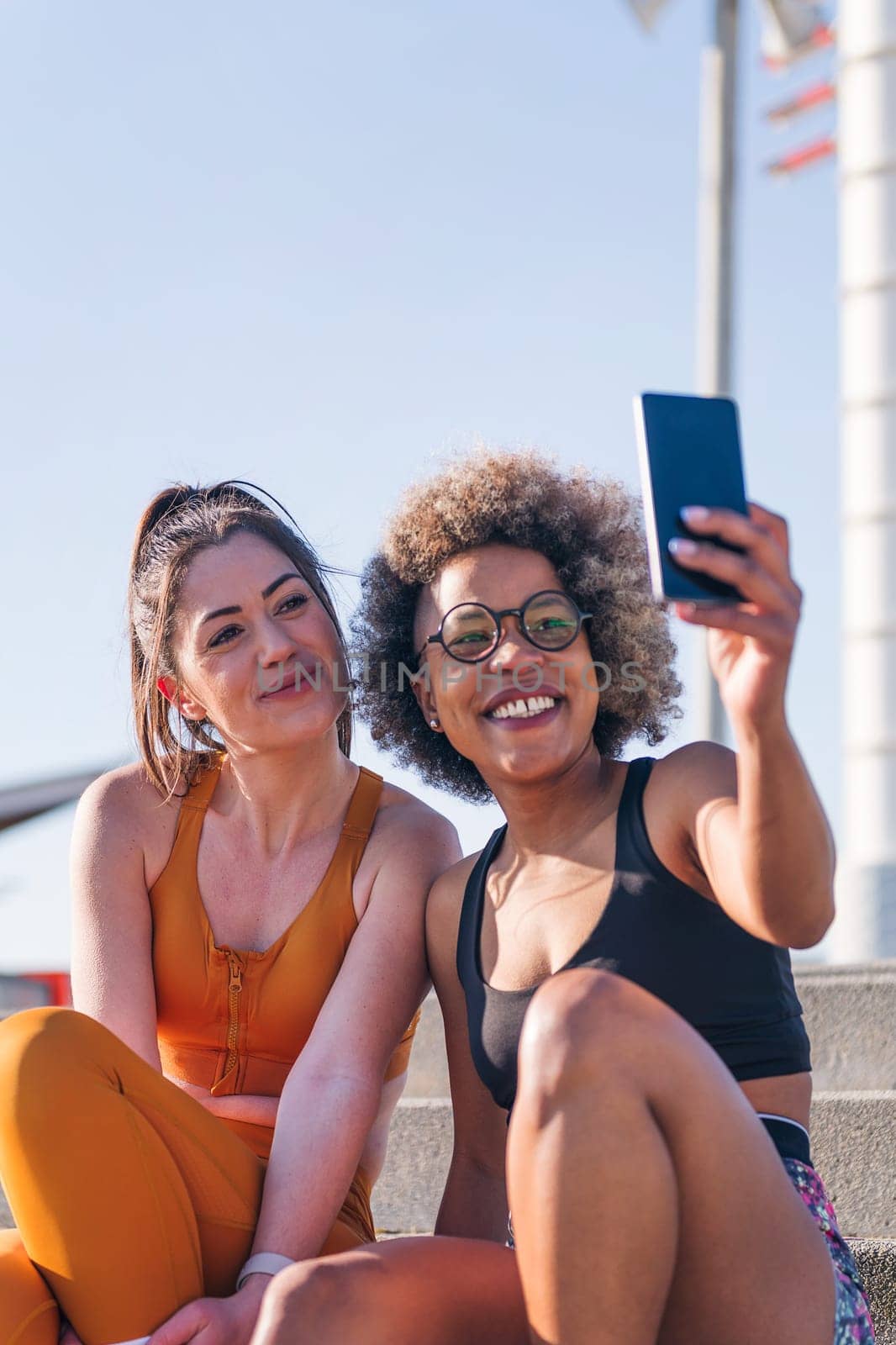 vertical photo of two female runner friends taking selfie photo with mobile phone sitting on urban stairs, concept of friendship and sporty lifestyle, copy space for text