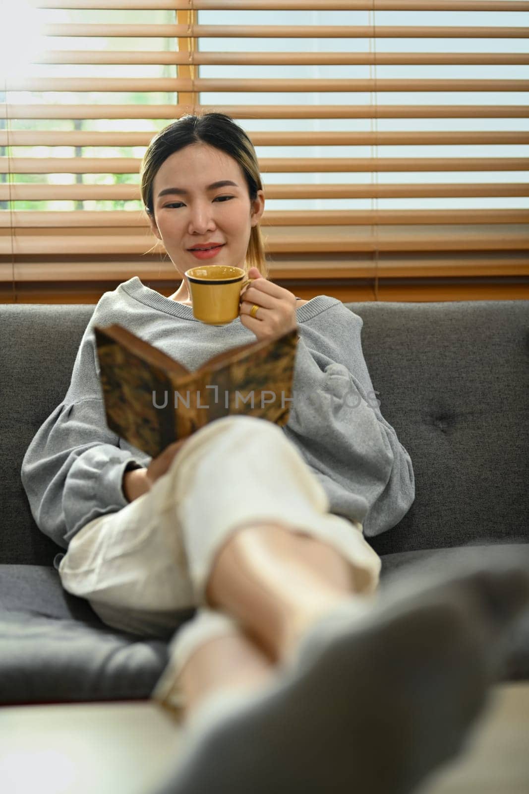 Relaxed young woman drinking hot coffee and reading book, spending free time in Sunday morning at home.