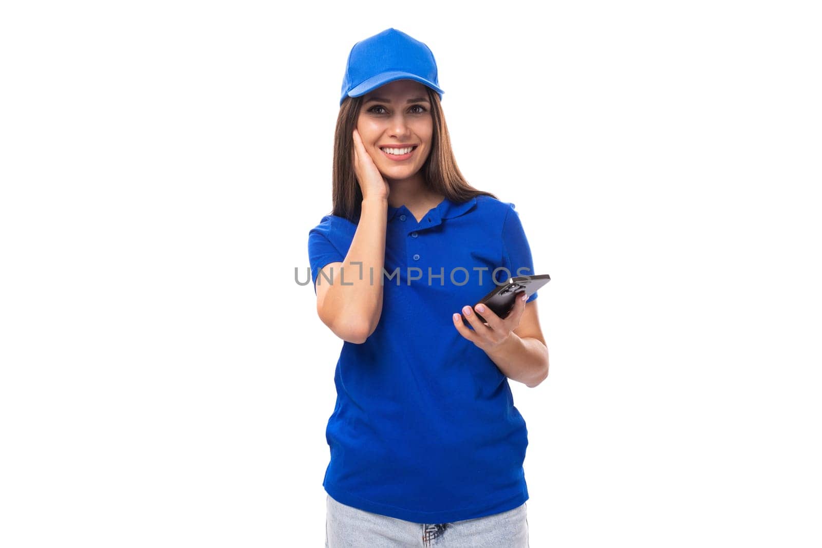 young brunette woman in a blue cap and t-shirt with a smartphone on a white background with copy space by TRMK