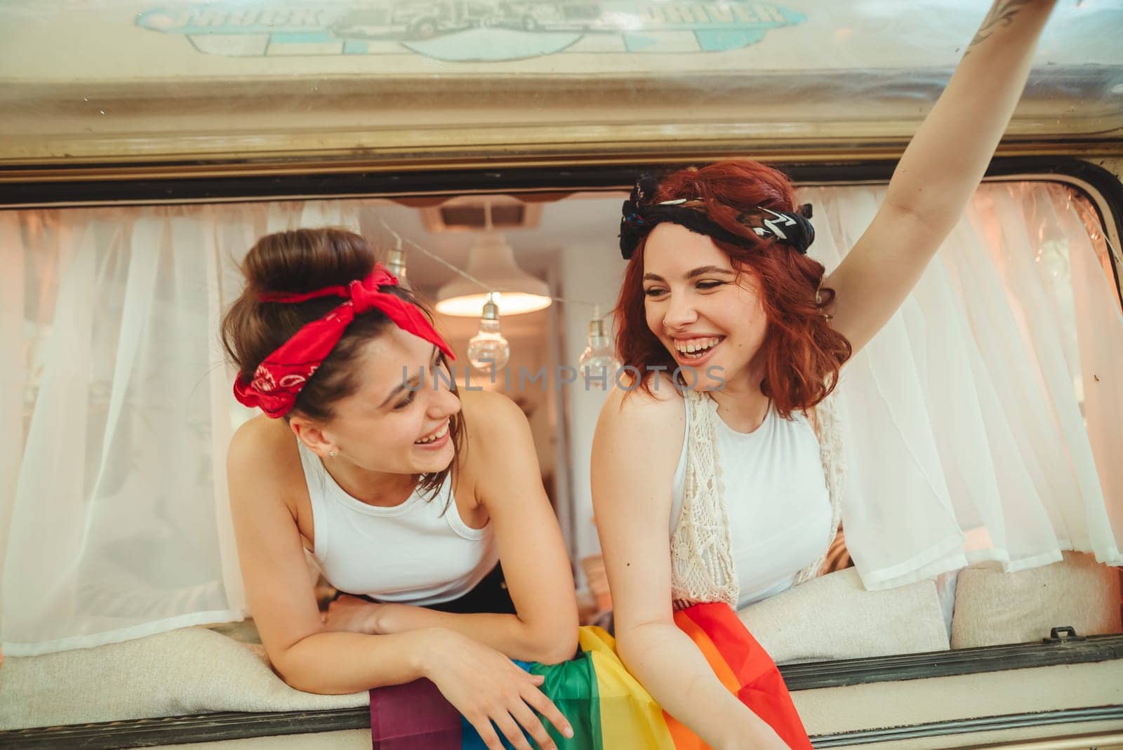 Portrait of a cute lesbian couple. Two girls spend time tenderly together watching movie on laptop in a camper trailer with LGBT flag on the wall. Love and attitude. LGBT concept. High quality photo