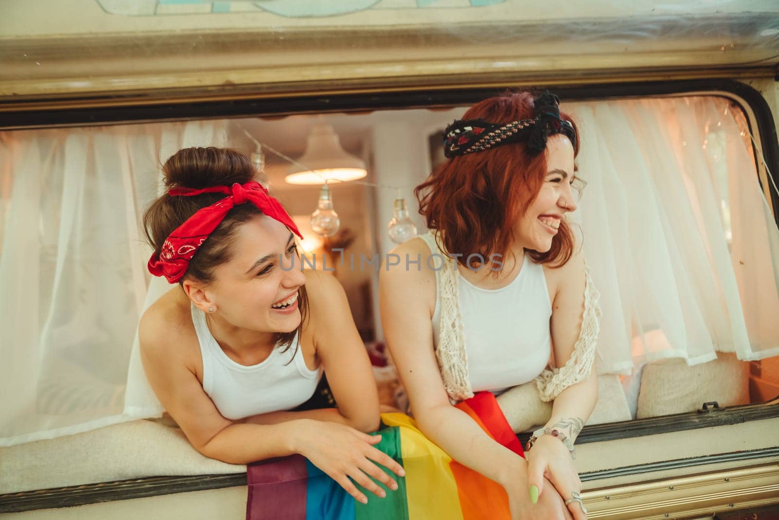 Portrait of a cute lesbian couple. Two girls spend time tenderly together watching movie on laptop in a camper trailer with LGBT flag on the wall. Love and attitude. LGBT concept. High quality photo