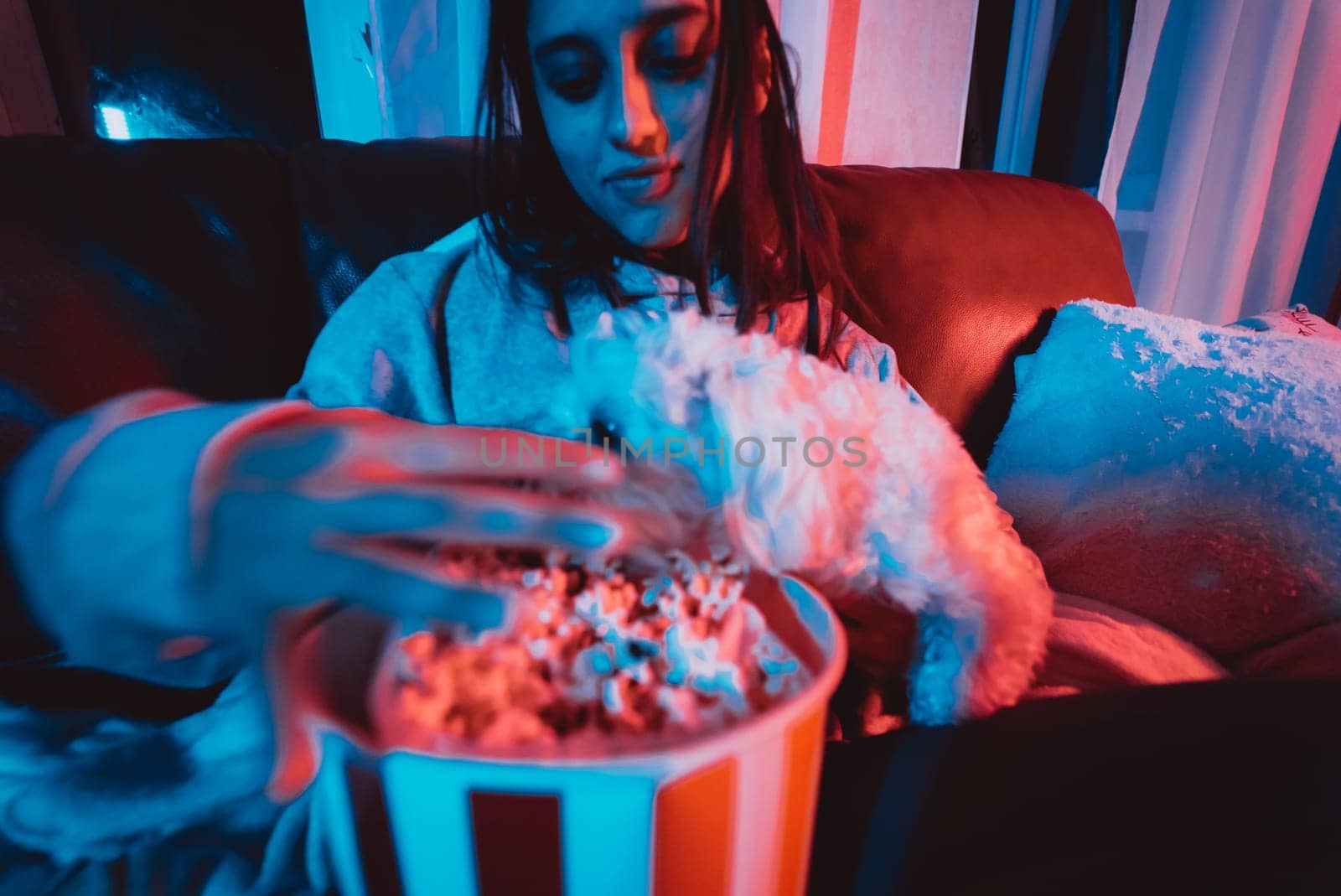 Close up portrait of Beautiful young girl at home in a dark room with a popcorn bucket playing with her dog and sits in front of a monitor or TV watching movie. Crazy emotions. High quality photo