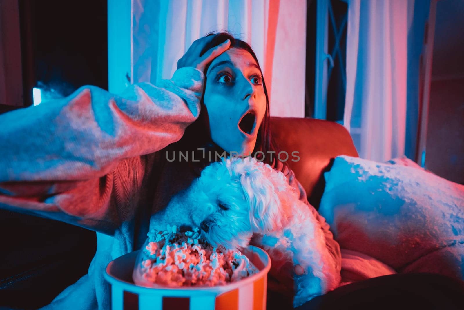 Close up portrait of Beautiful young girl at home in a dark room with a popcorn bucket playing with her dog and sits in front of a monitor or TV watching movie. Crazy emotions. High quality photo