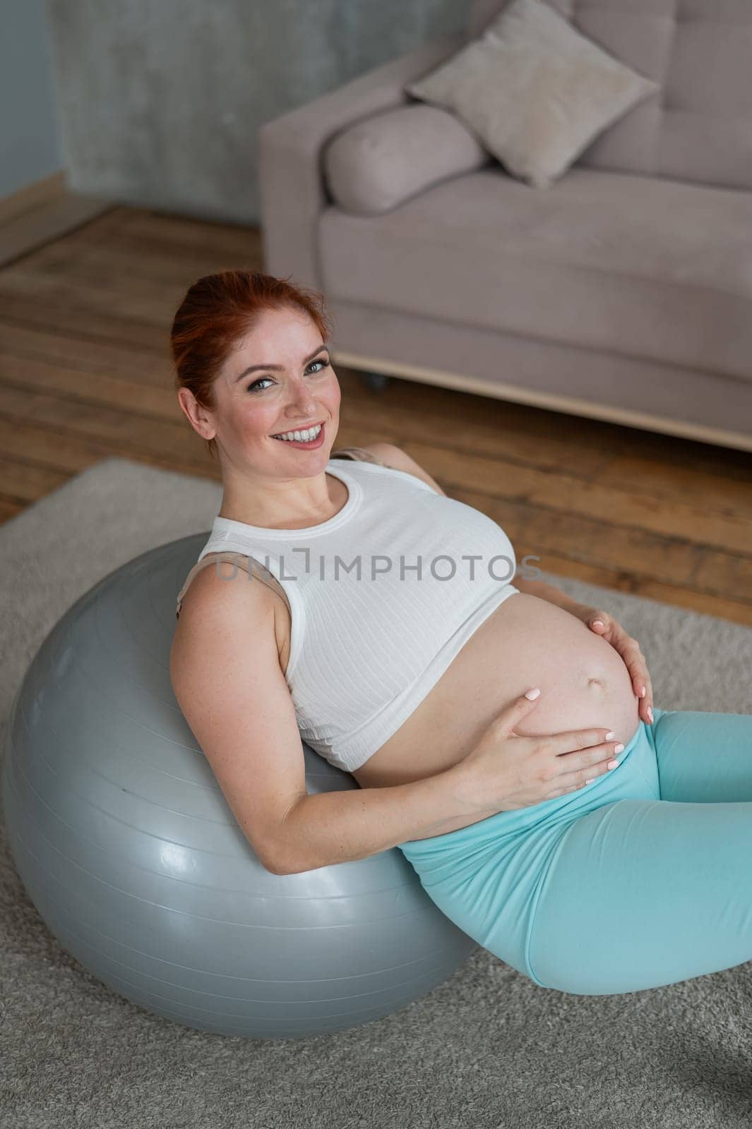 Happy pregnant woman lies with her back on a fitball and keeps her hands on her tummy at home
