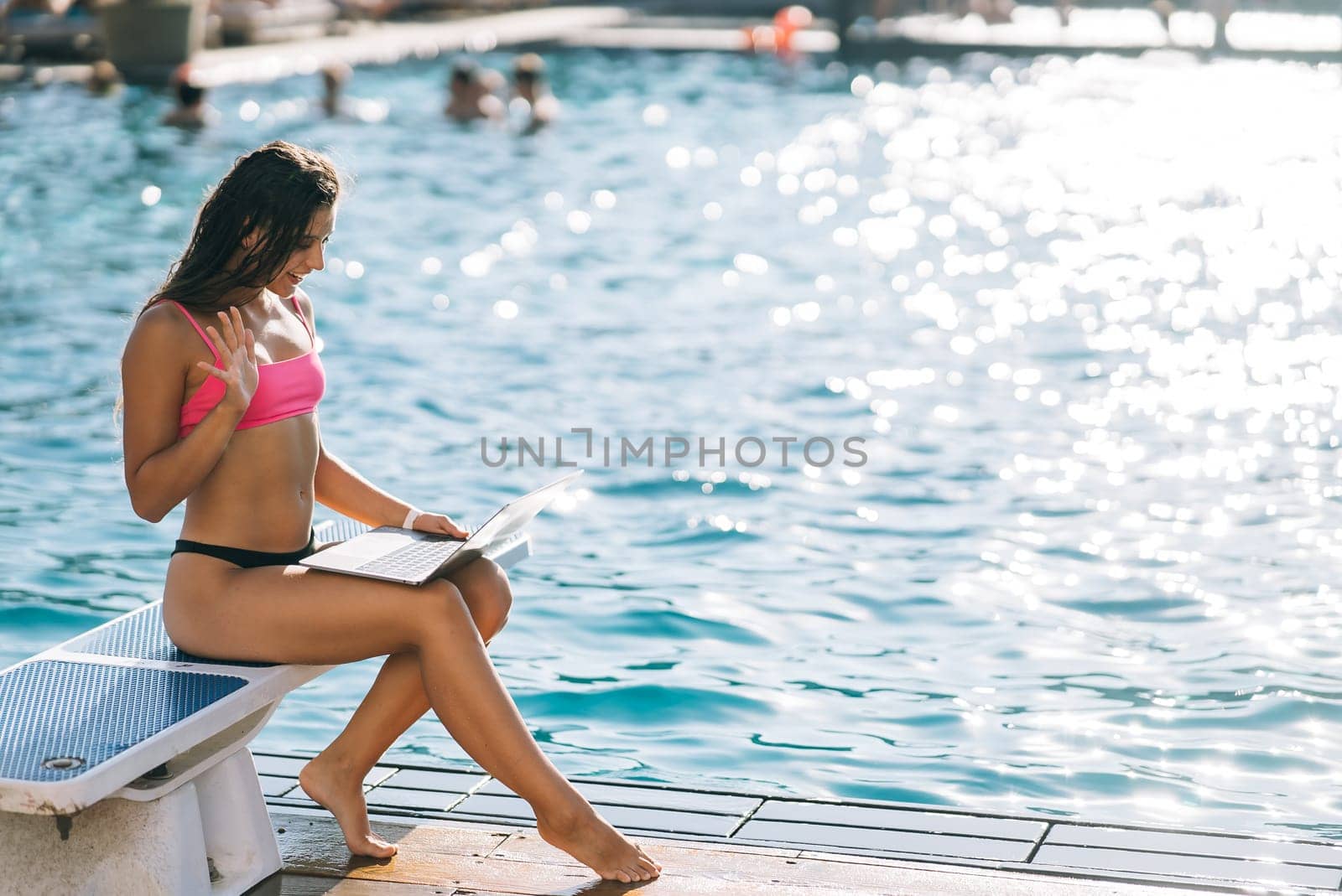 Cute young woman sunbathing sitting in front of the pool, working with a laptop at summer day
