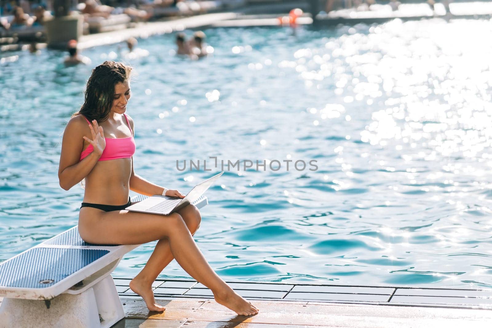 Cute young woman sunbathing sitting in front of the pool, working with a laptop at summer day