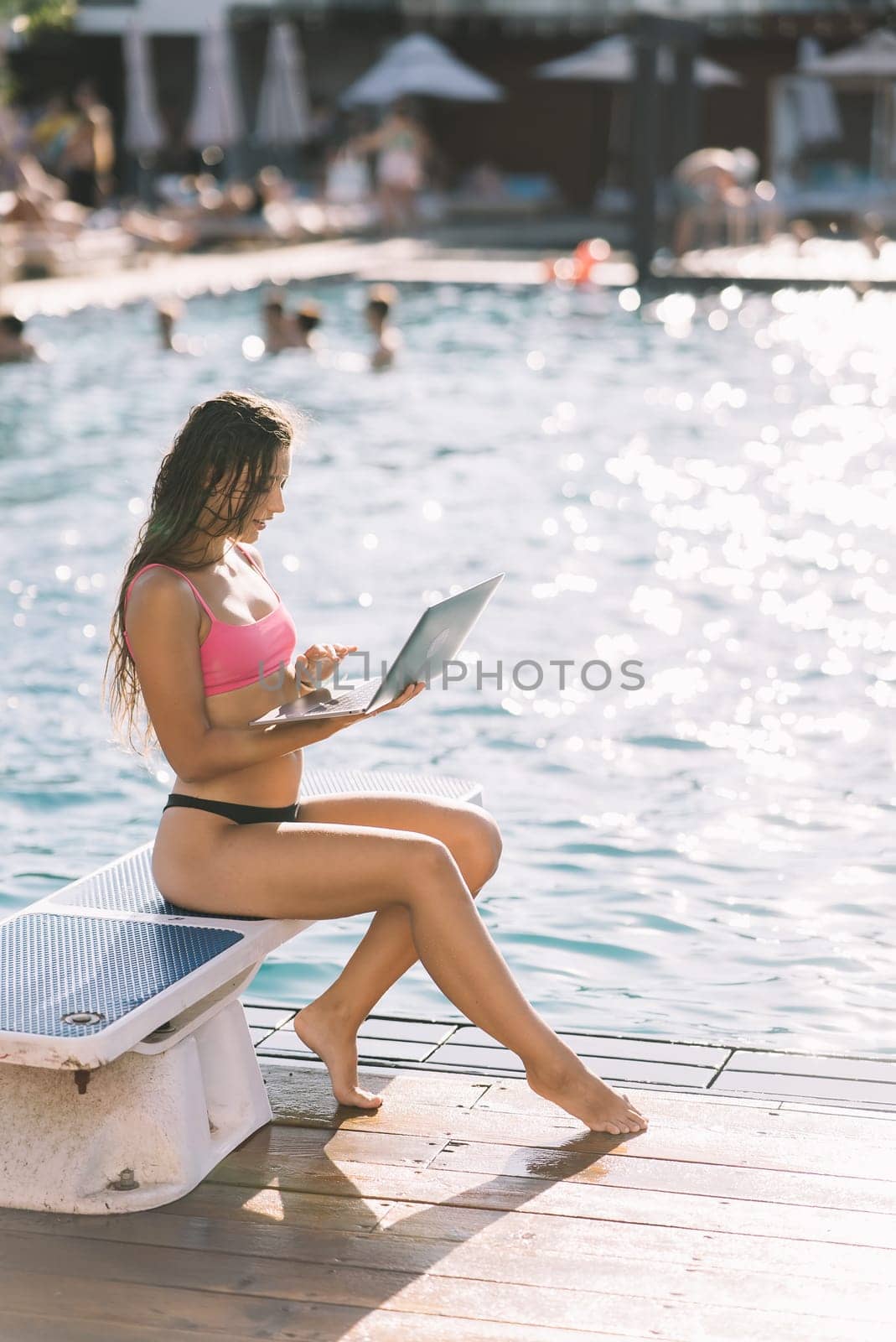 Cute young woman sitting in front of the pool, working with a laptop at summer day