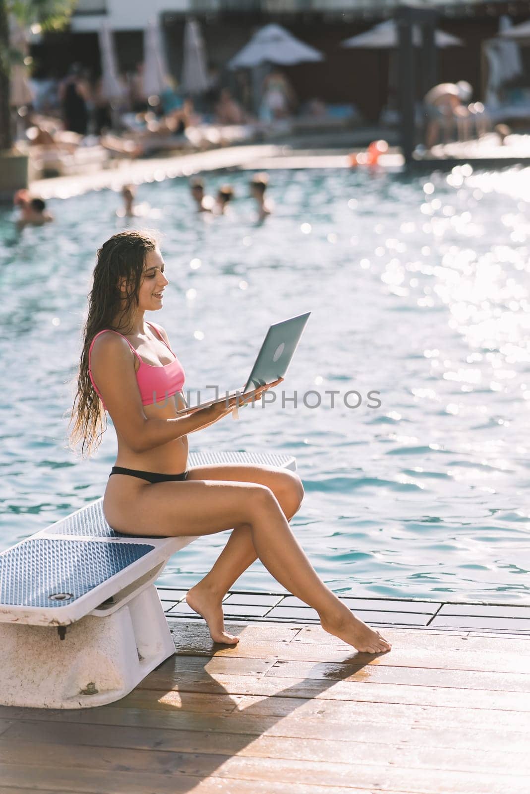 Cute young woman sitting in front of the pool, working with a laptop at summer day
