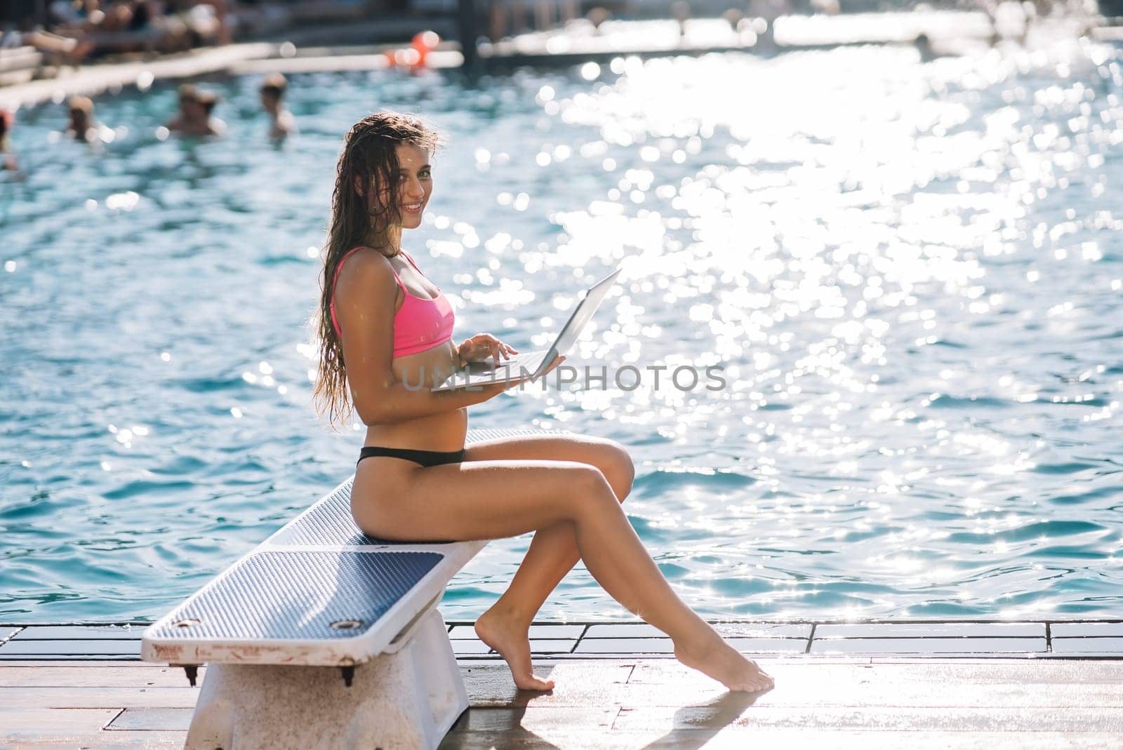 Cute young woman sitting in front of the pool, working with a laptop at summer day