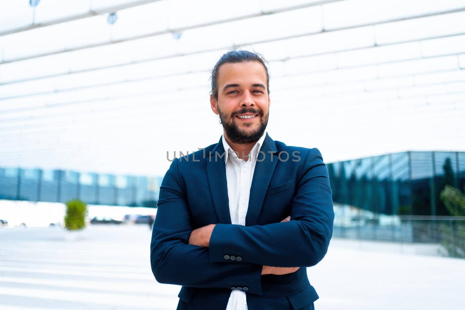 Confidence successful businessman or worker in suit with beard standing business district glass building arm crossed looking at camera and smile. Hispanic male business person portrait. Copy space
