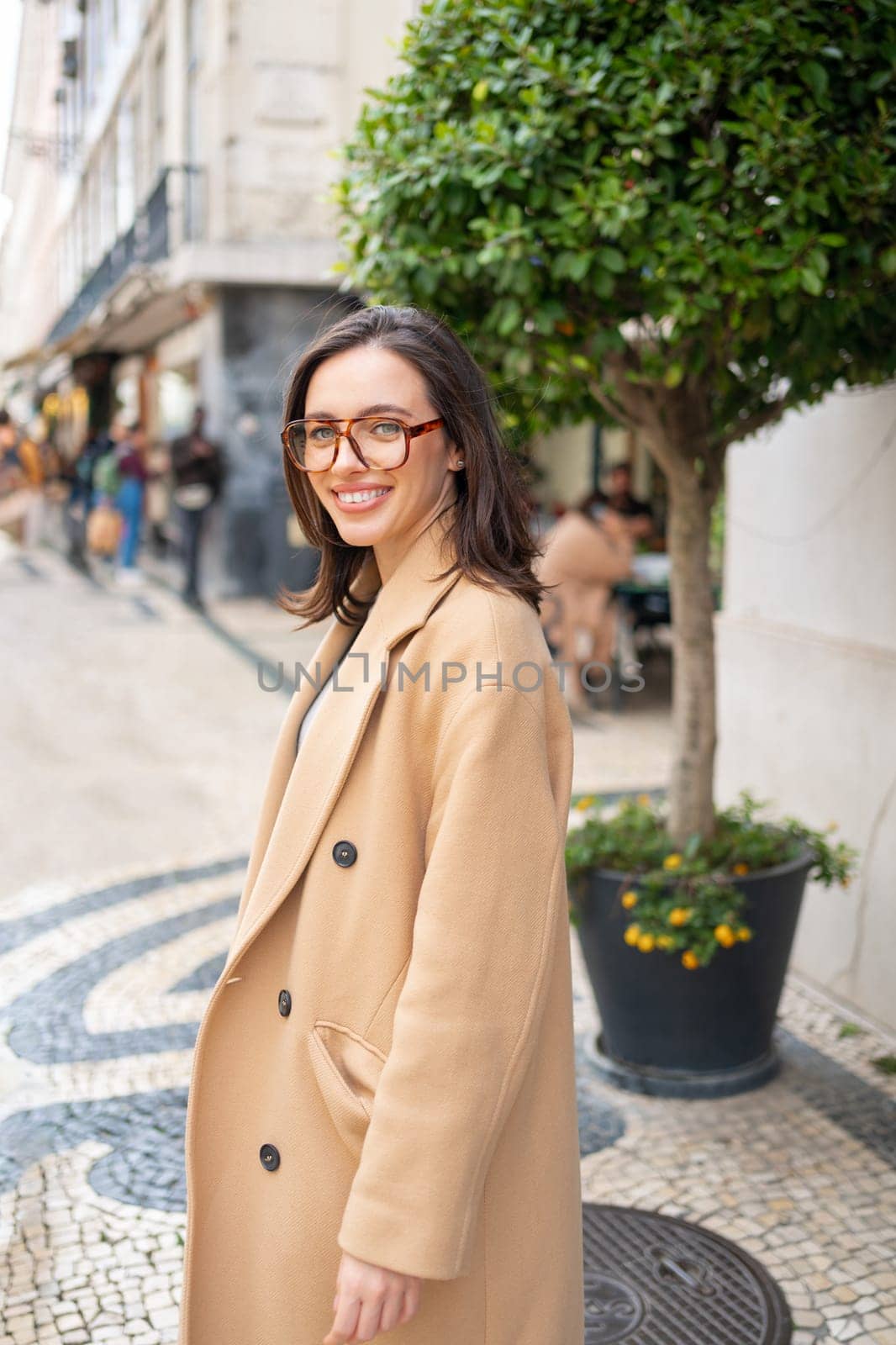 Friendly smiling attractive woman leaving building on background green hedge in the street of European city. Happy confident woman in glasses dressed stylish trench coat smiling looking at camera