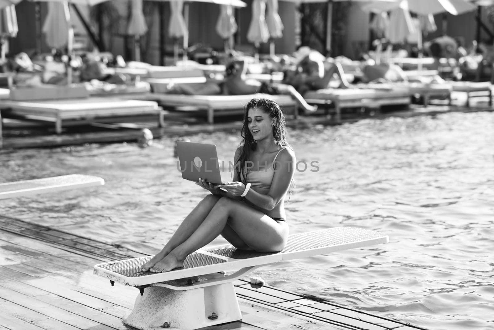 Cute young woman sunbathing sitting in front of the pool, working with a laptop at summer day. Black and white photo