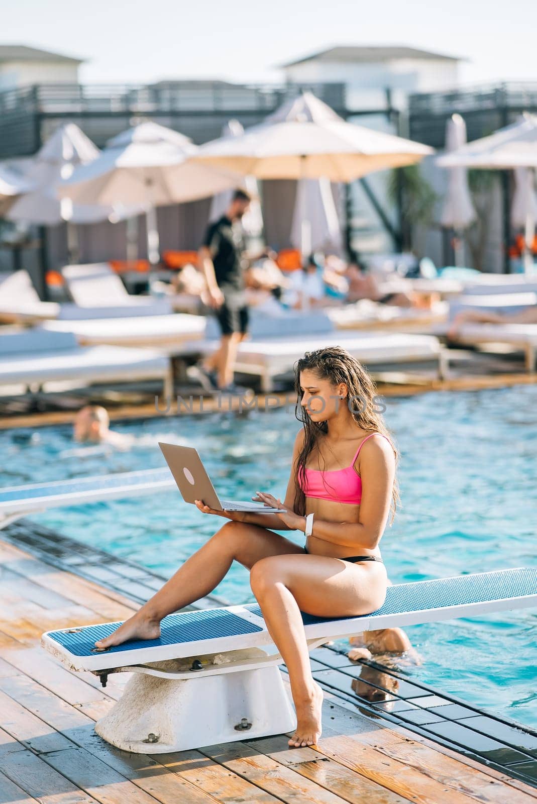 Cute young woman sunbathing sitting in front of the pool, working with a laptop at summer day