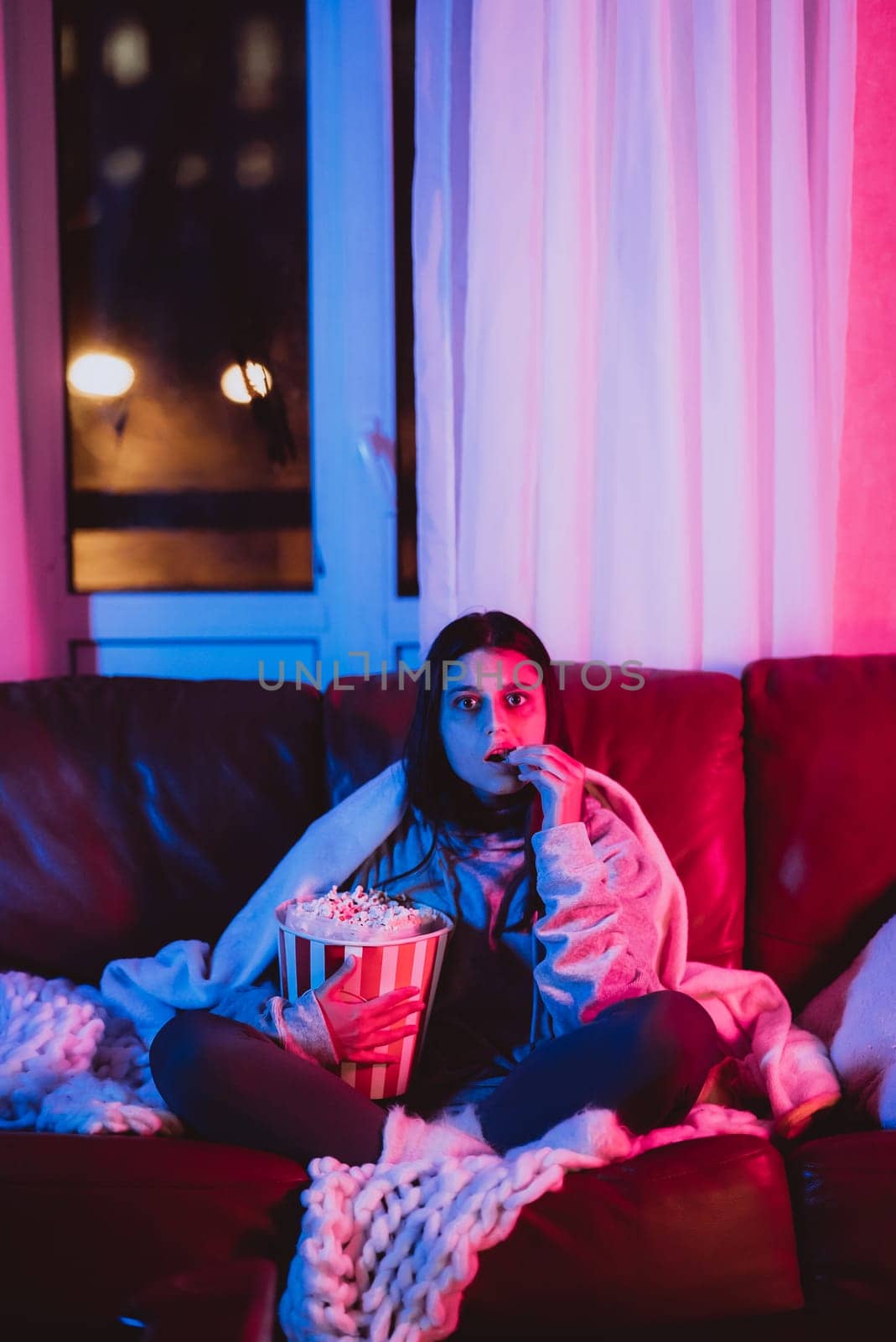 Beautiful young girl eating popcorn watching movie at home in a dark room with a popcorn bucket and playing with her dog and sits in front of a monitor or TV watching movie. Crazy emotions. High quality photo
