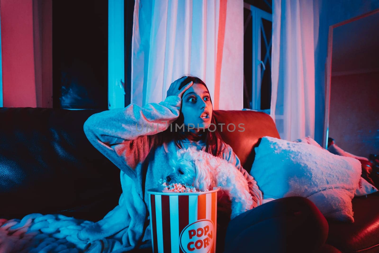 Beautiful young girl at home in a dark room with a popcorn bucket playing with her dog and sits in front of a monitor or TV watching movie. High quality photo