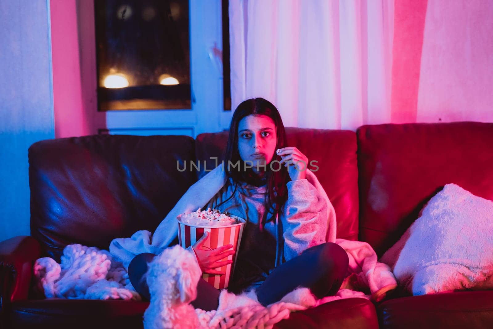 Beautiful young girl eating popcorn watching movie at home in a dark room with a popcorn bucket and playing with her dog and sits in front of a monitor or TV watching movie. Crazy emotions. High quality photo