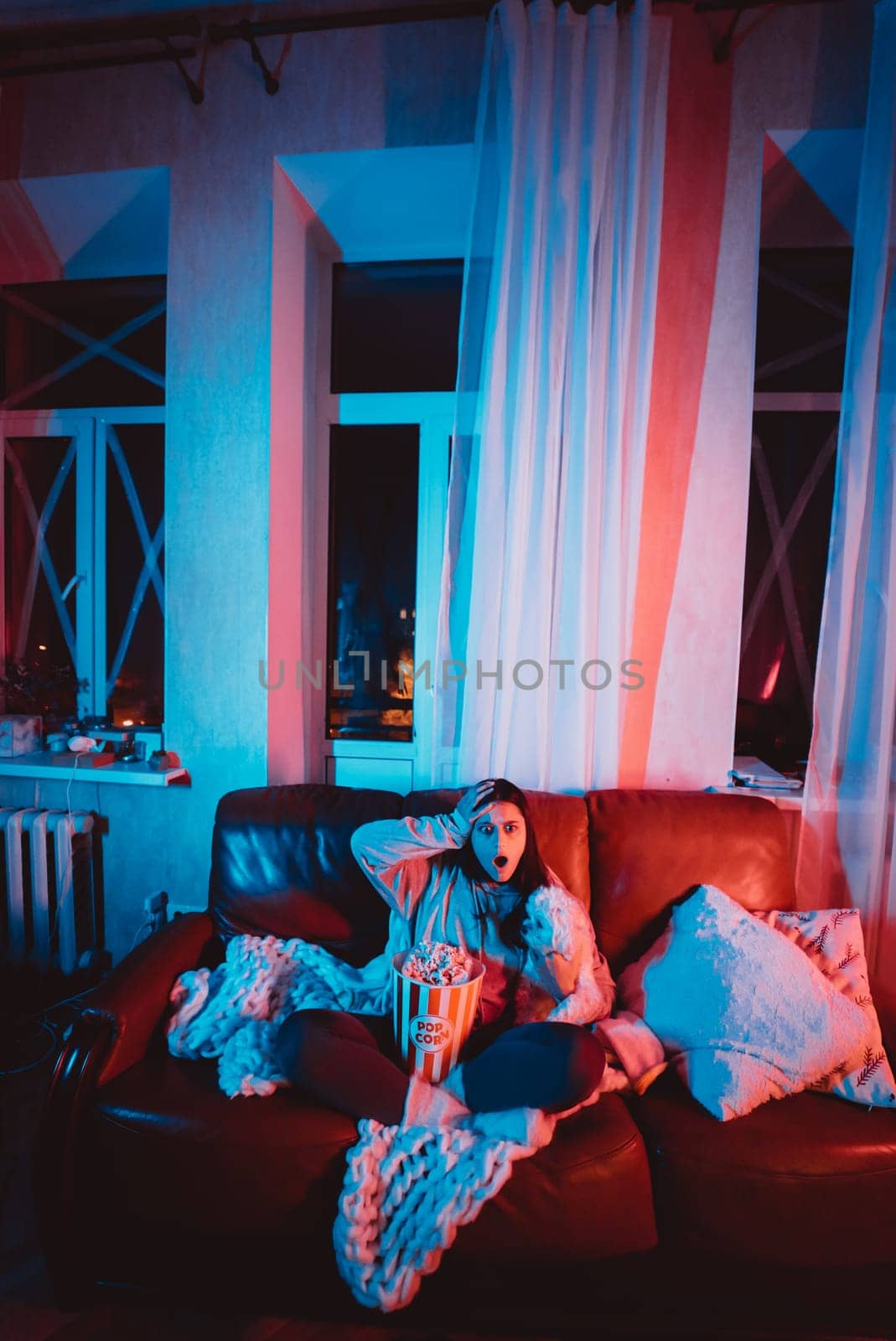 Beautiful young girl watching movie at home in a dark room with a popcorn bucket playing with her dog and sits in front of a monitor or TV watching movie. Crazy emotions. High quality photo