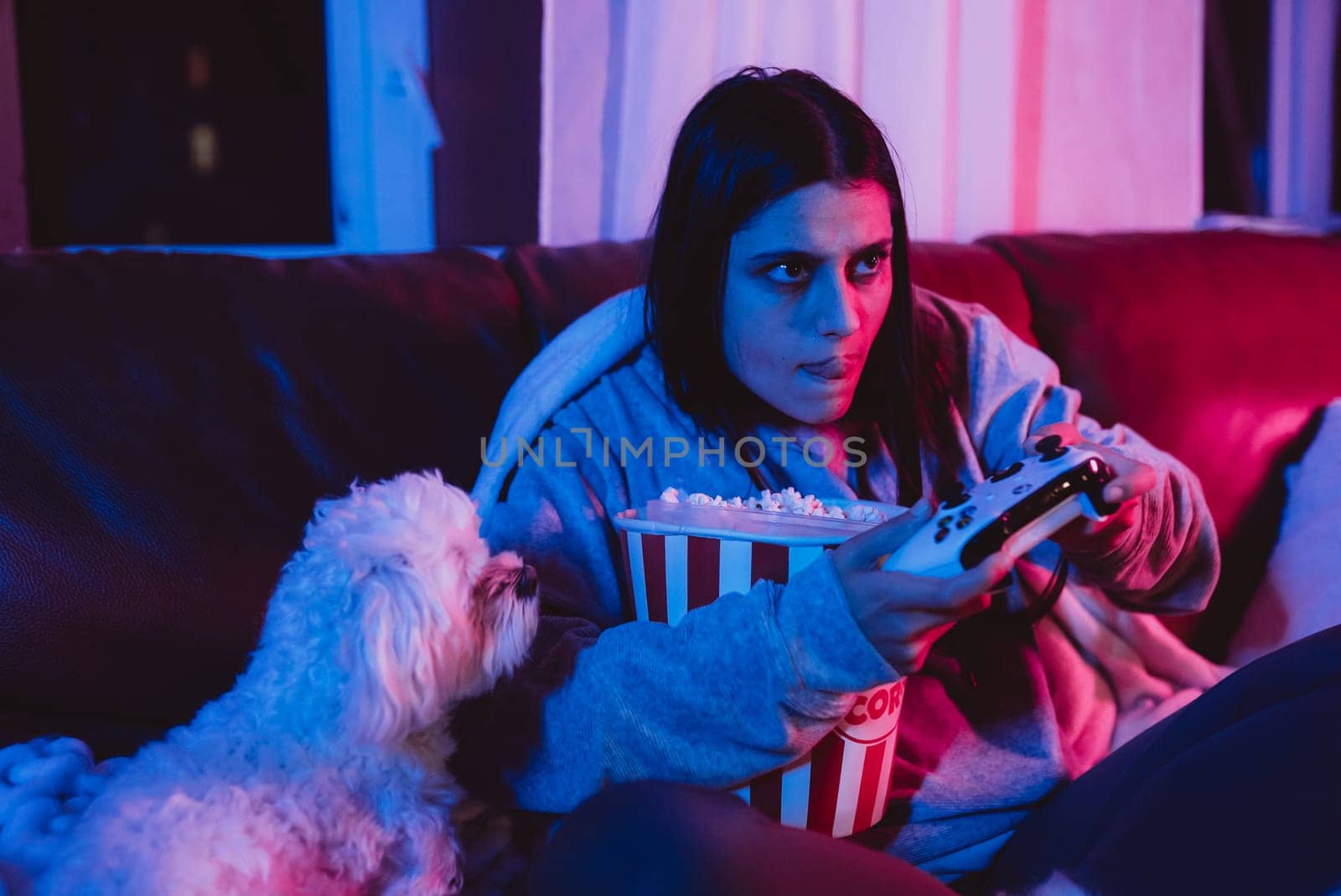A gamer or a streamer girl at home in a dark room with a game controller and popcorn bucket playing with her dog and sits in front of a monitor or TV. High quality photo