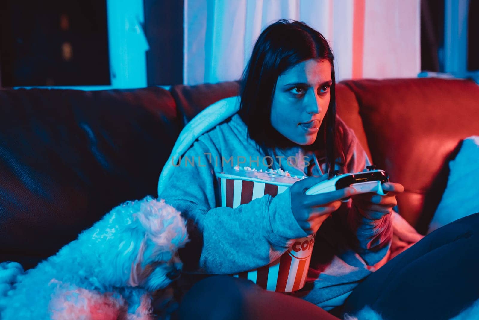 A gamer or a streamer girl at home in a dark room with a game controller and popcorn bucket playing with her dog and sits in front of a monitor or TV. High quality photo