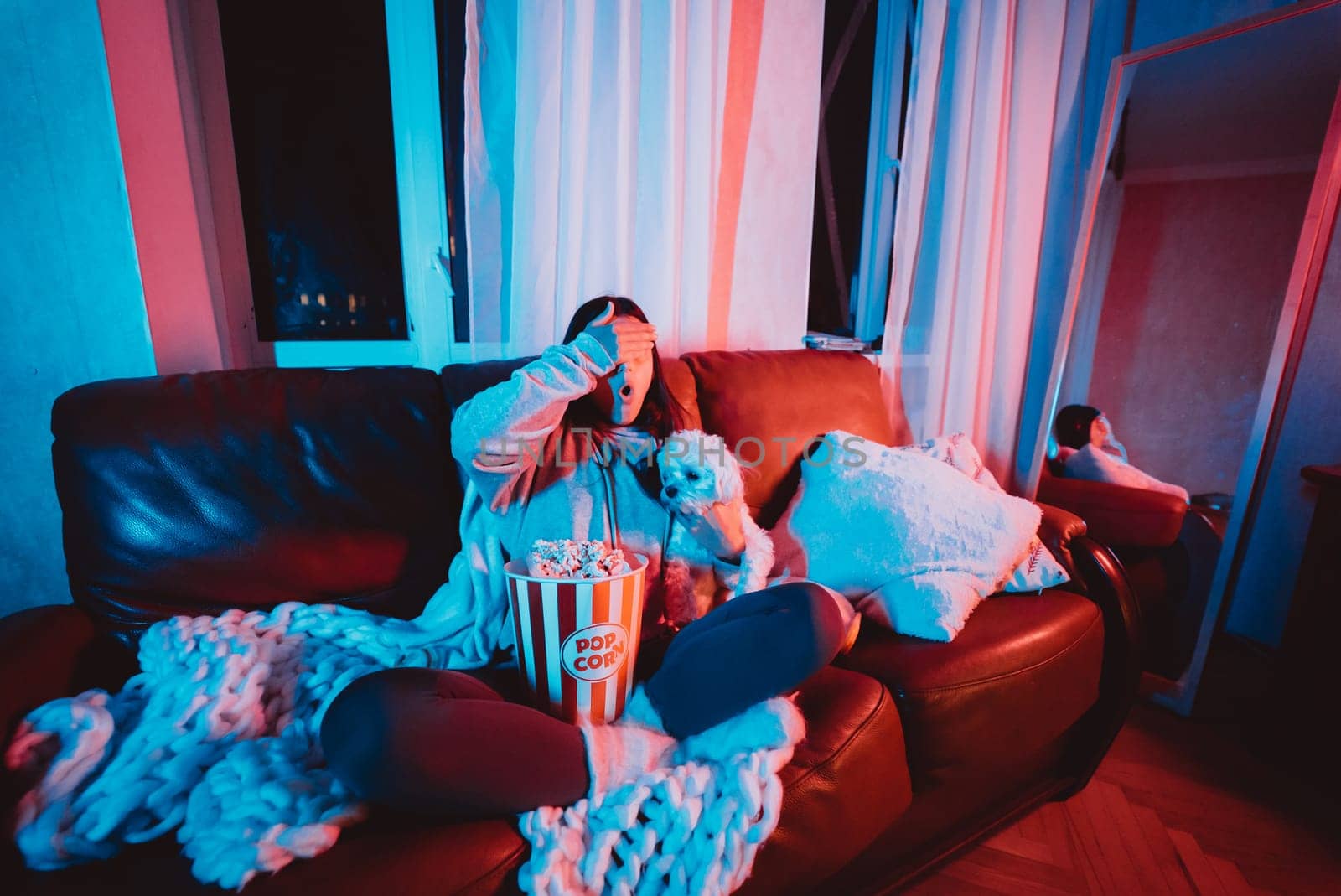Close up portrait of Beautiful young girl hide her eyes watching movie at home in a dark room with a popcorn bucket playing with her dog and sits in front of a monitor or TV watching movie. Crazy emotions. High quality photo