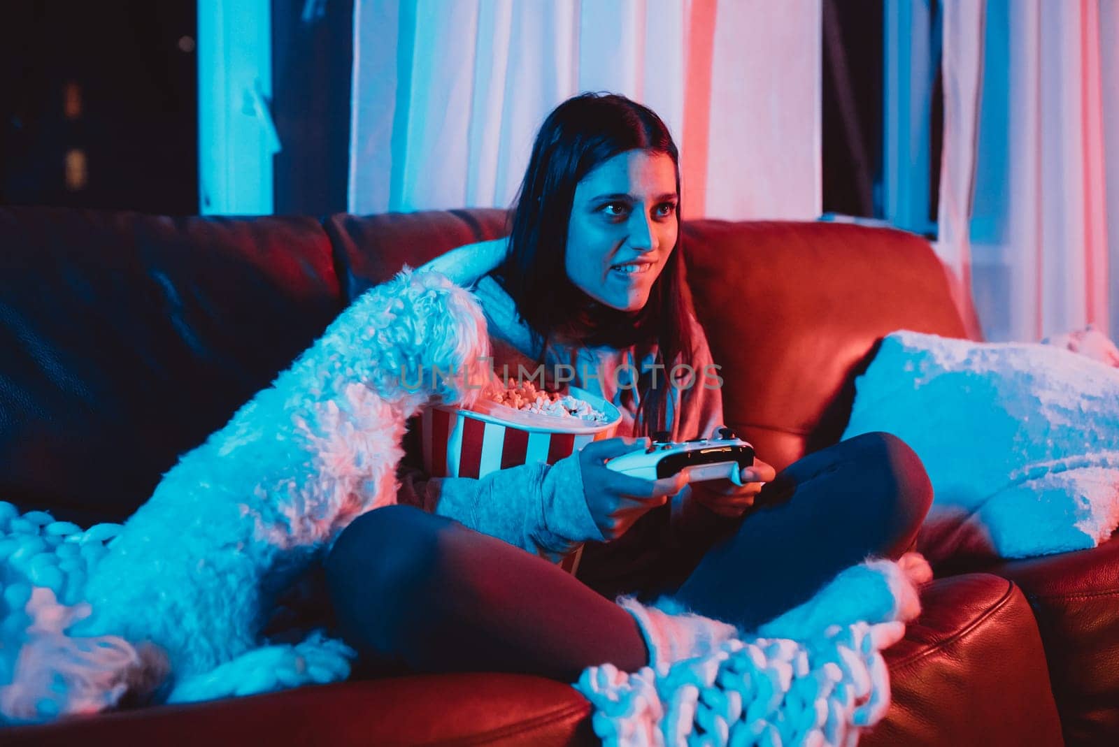 A gamer or a streamer girl at home in a dark room with a game controller and popcorn bucket playing with her dog and sits in front of a monitor or TV. High quality photo