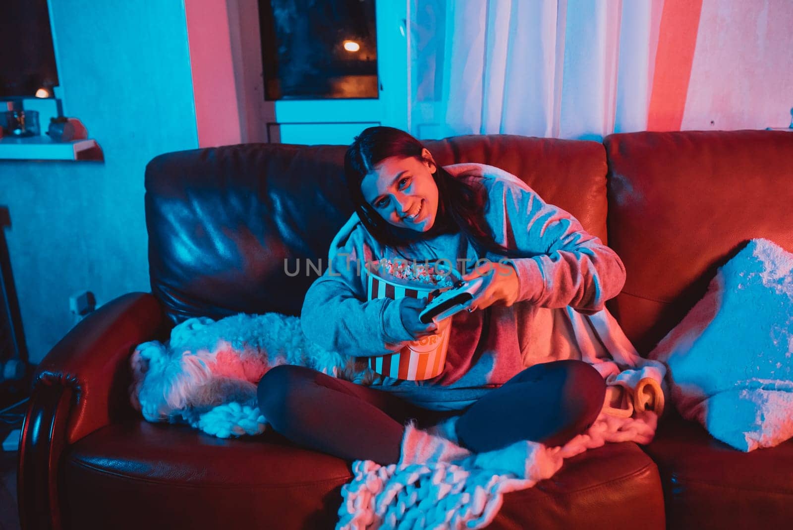 A gamer or a streamer girl at home in a dark room with a game controller and popcorn bucket playing with her dog and sits in front of a monitor or TV. High quality photo