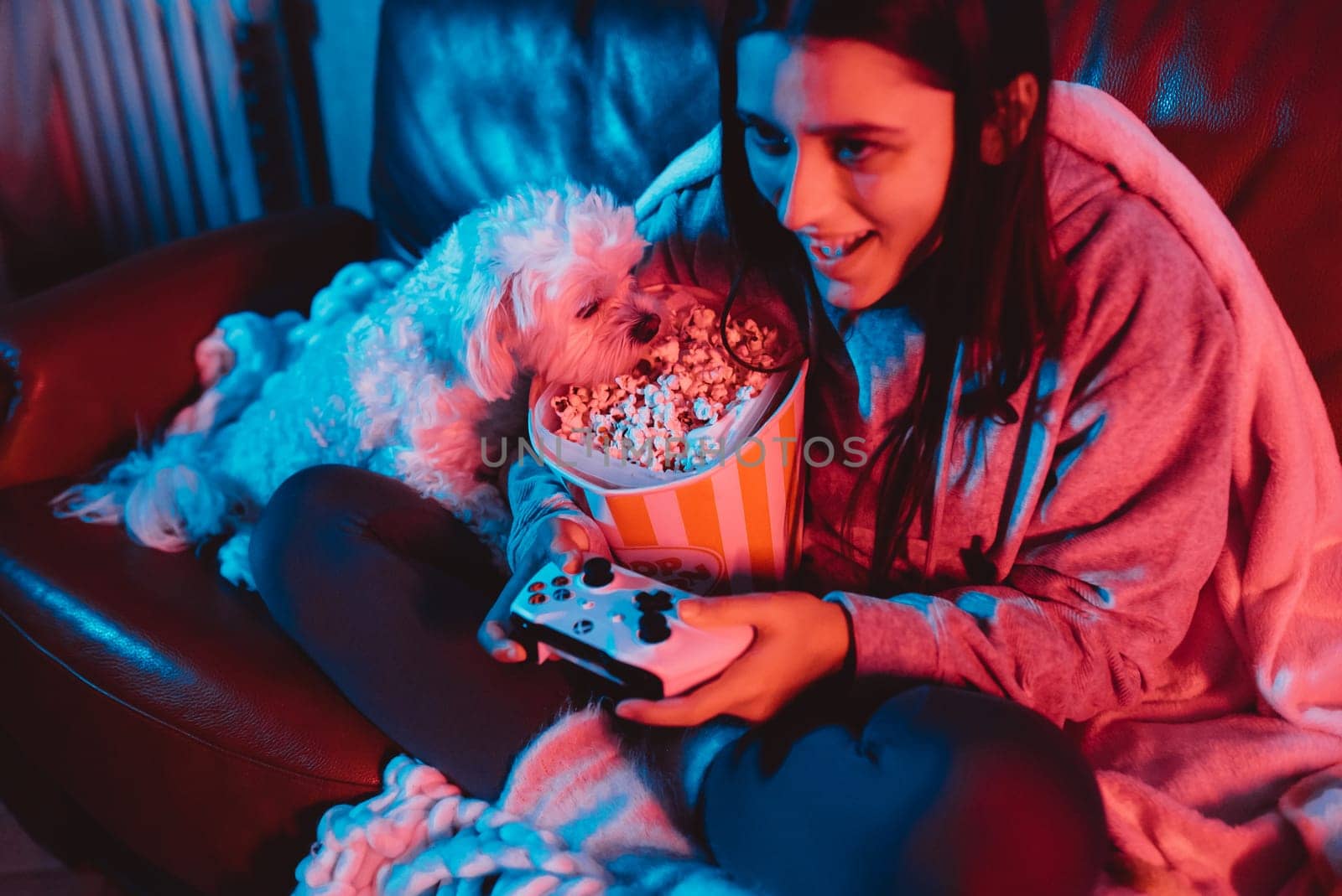 A gamer or a streamer girl at home in a dark room with a game controller and popcorn bucket playing with her dog and sits in front of a monitor or TV. High quality photo
