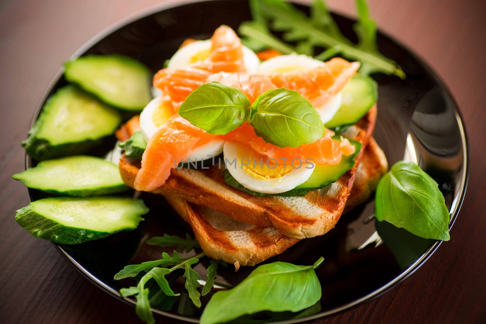 Cooked sandwich with lightly salted red fish, cucumbers and boiled eggs with herbs, in a plate.