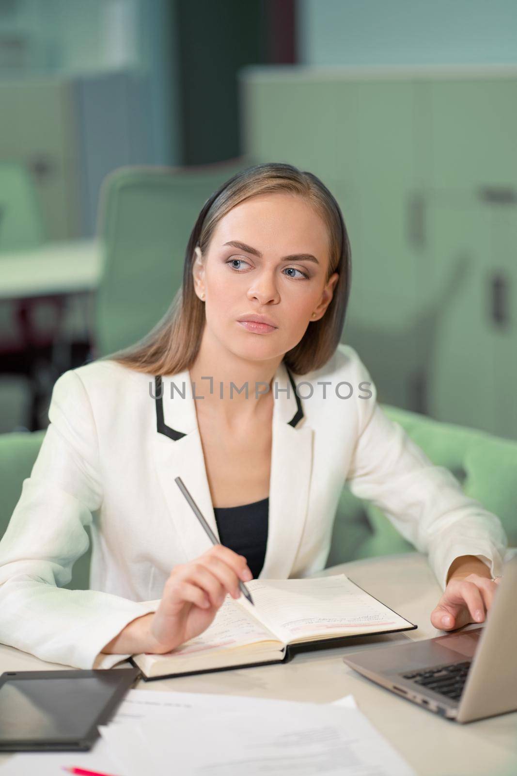 Beautiful businesswoman in white jacket making notes in her journal holding a pen her hand sitting in front of laptop. Office worker looking at her working place.