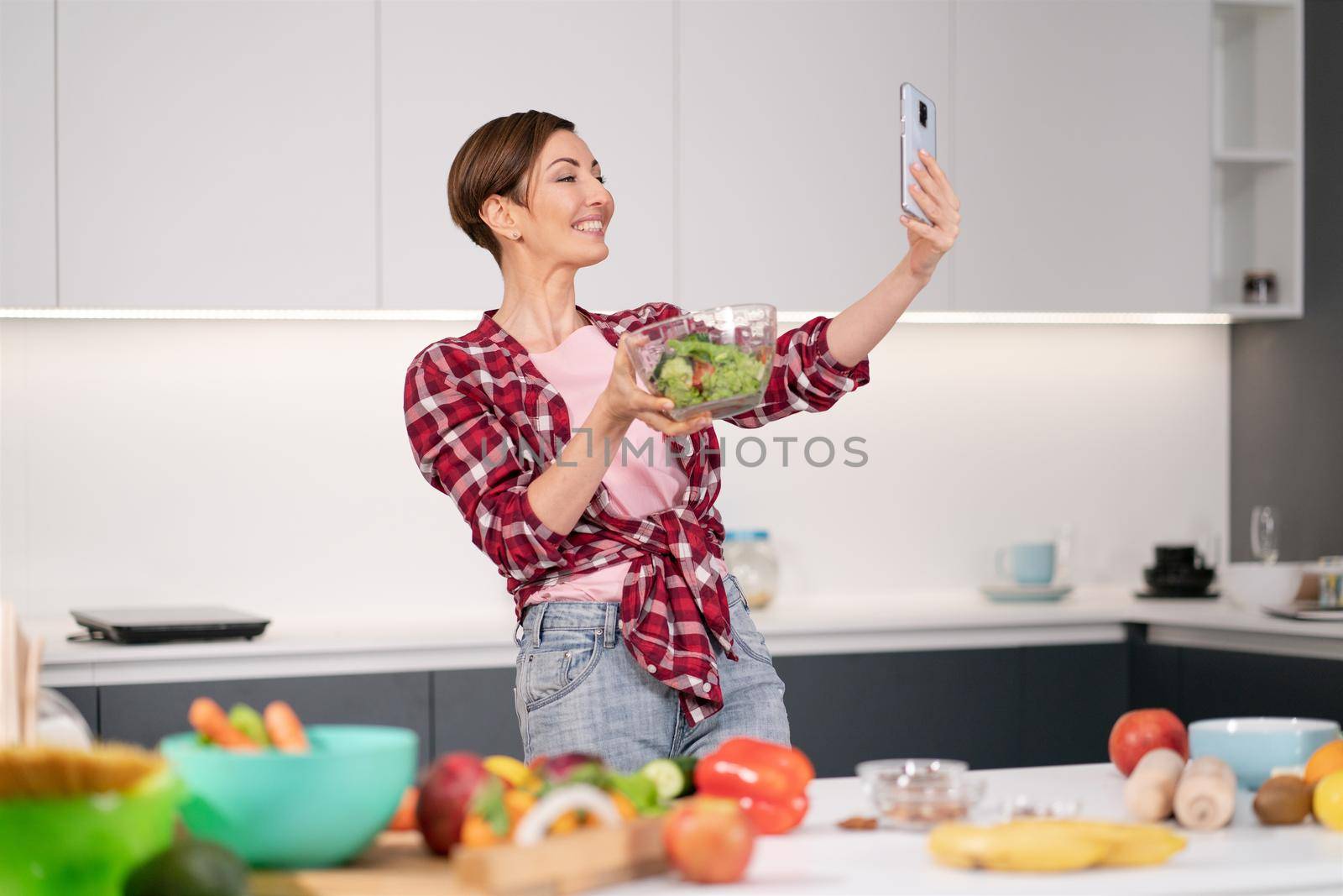 Pretty woman happy taking selfie using her smartphone while cooking fresh salad wearing a plaid shirt with a bob hair style. Healthy food leaving - vegan concept by LipikStockMedia