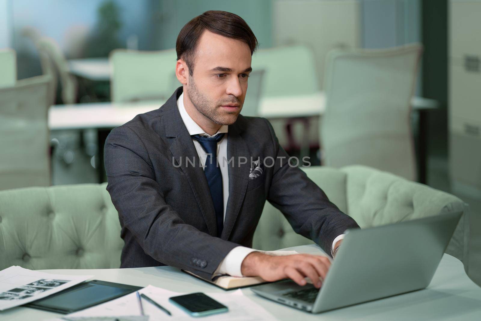 Freelancer sitting in front laptop in bright coworking space. Handsome man in business suit working on laptop, freelancer job in progress.